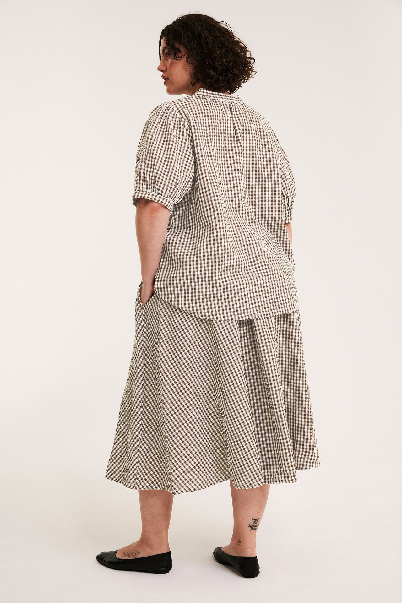 Kvinne sett bakfra/siden i et brunt rutete vidt seersucker-skjørt og en matchende topp.