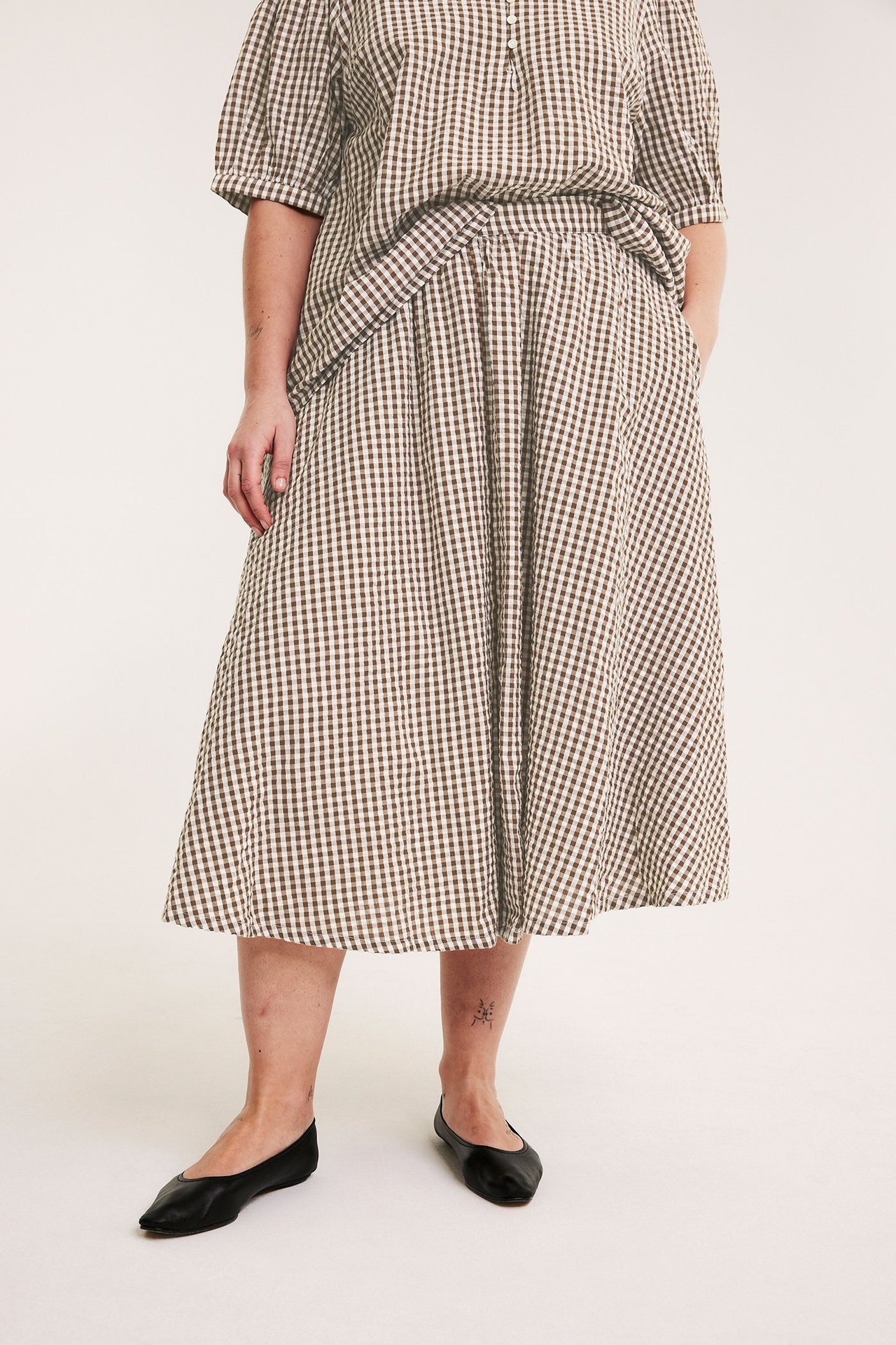 Edestä, nainen leveässä ruskearuudullisessa seersucker-hameessa, yhteensopiva toppi ja mustat ballerinat.
