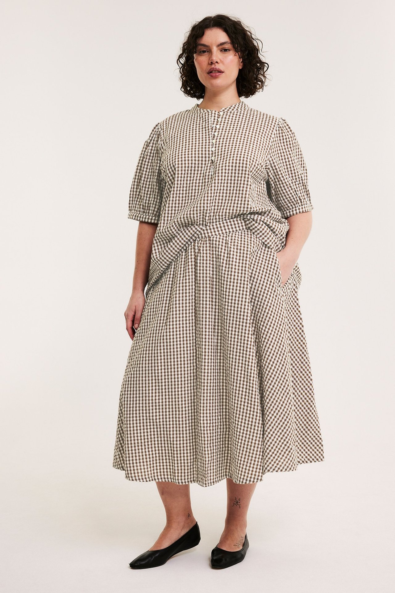 Kobieta z przodu w brązowej spódnicy midi w kratę z seersuckeru z kieszeniami i pasującej bluzce.