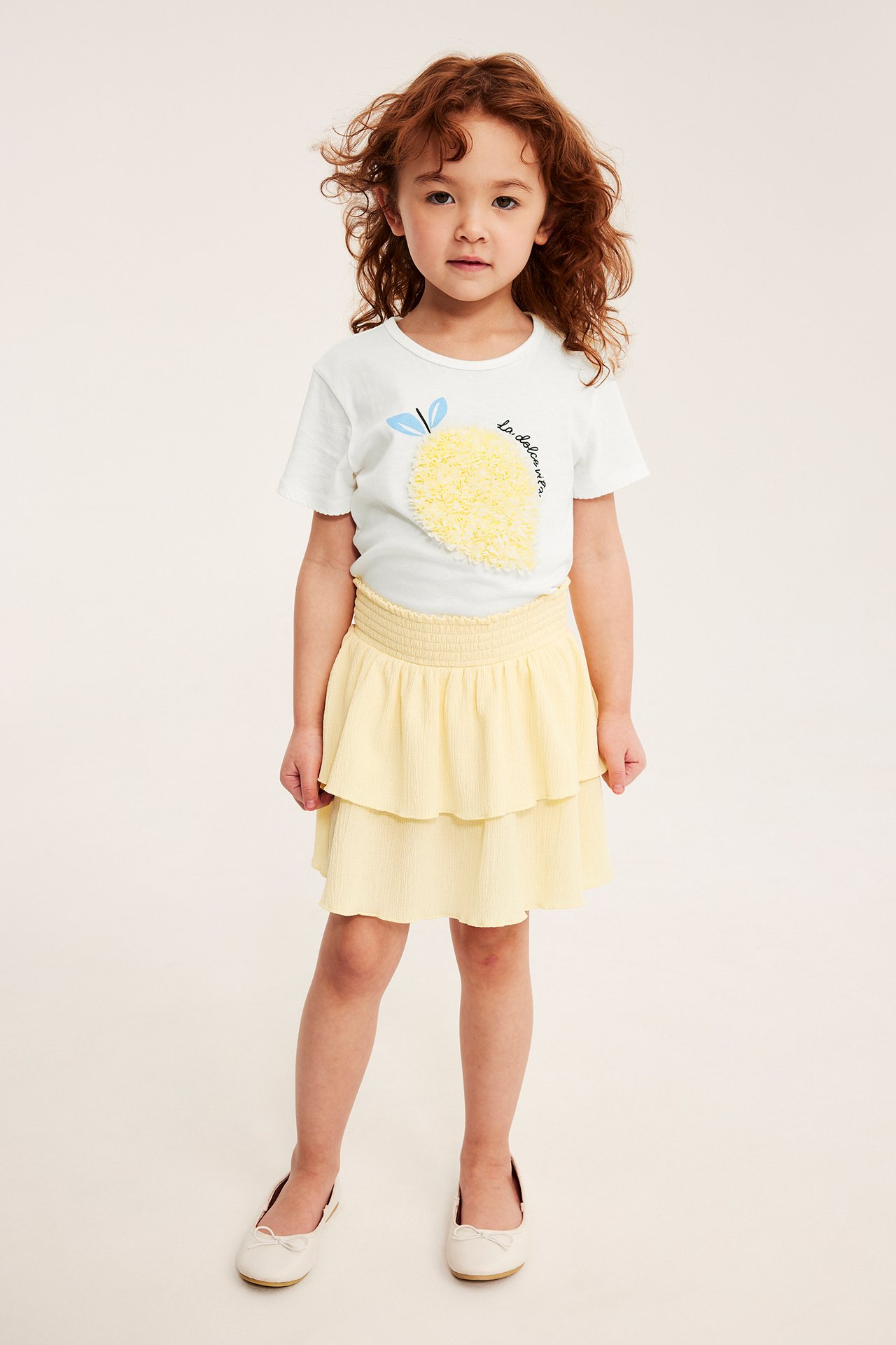 Edestä kuvattu lapsi yllään luonnonvalkoinen puuvillatoppi keltaisella sitruuna-applikaatiolla, keltainen kerroshame ja valkoiset ballerinakengät.