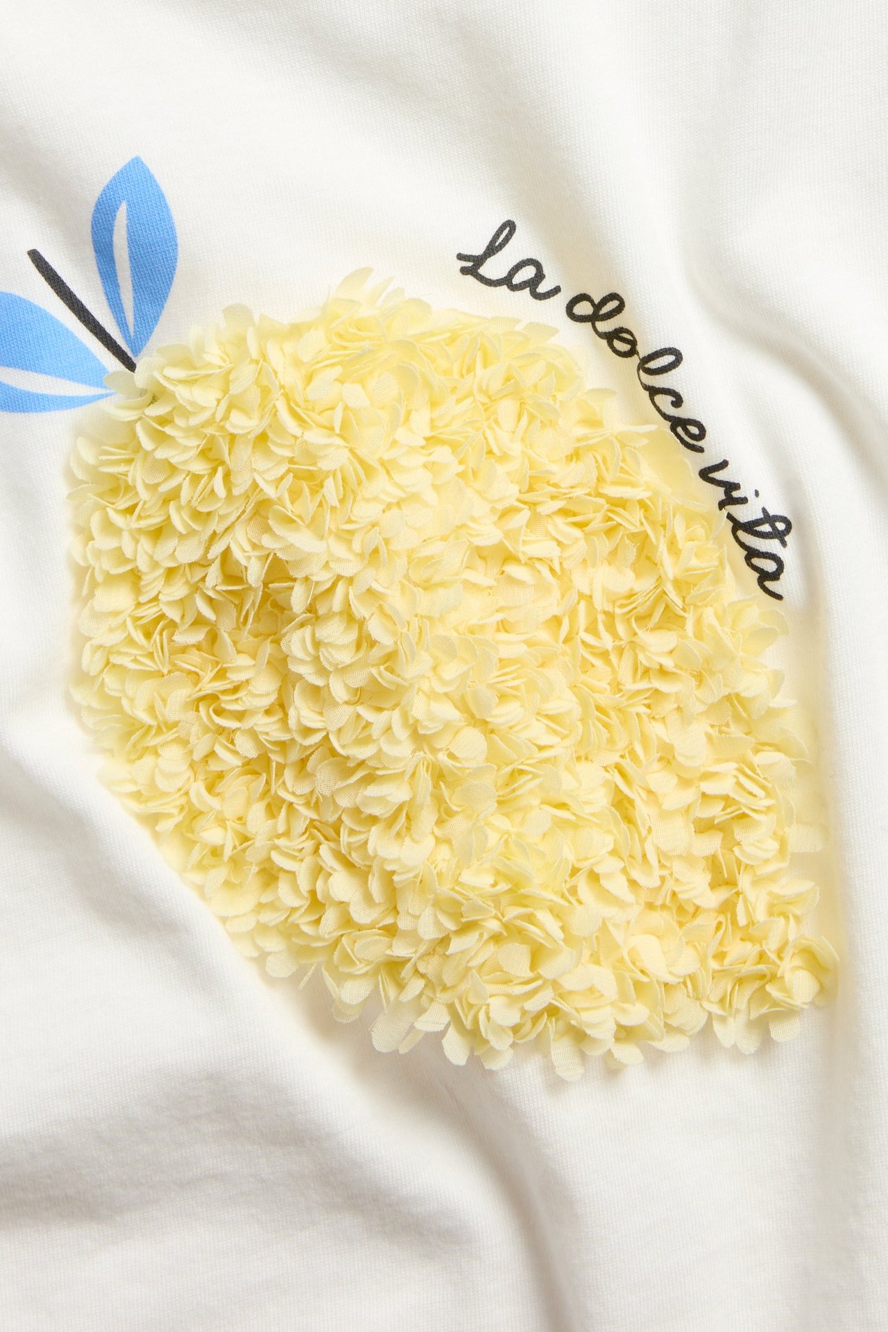 Zbliżenie na ecru bawełniany top z żółtą aplikacją 3D cytryny i czarnym napisem 'la dolce vita'.