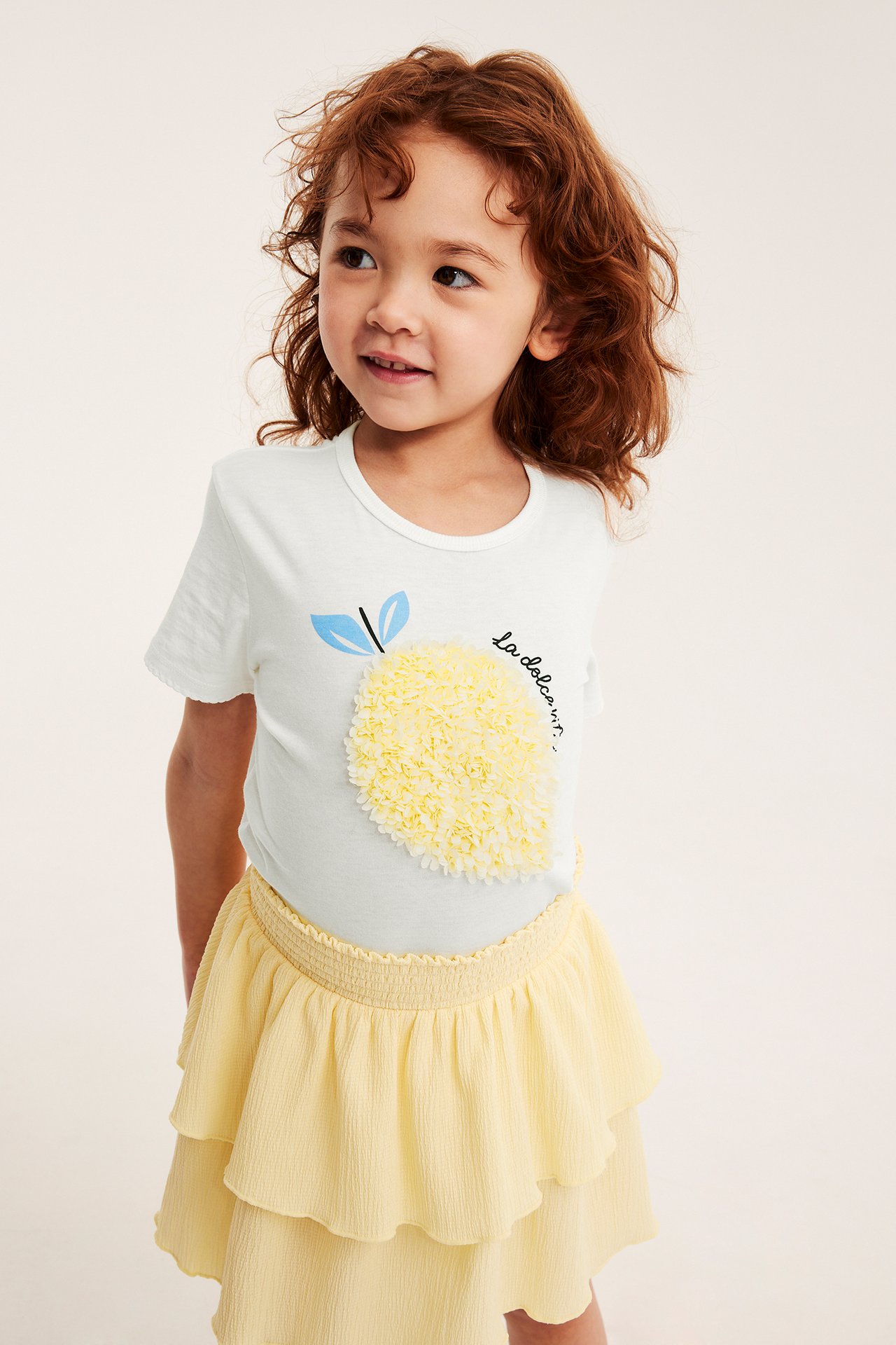 Widok z przodu: Dziecko w kremowym bawełnianym topie z żółtą aplikacją cytryny i jasnożółtej spódniczce z falbanami.