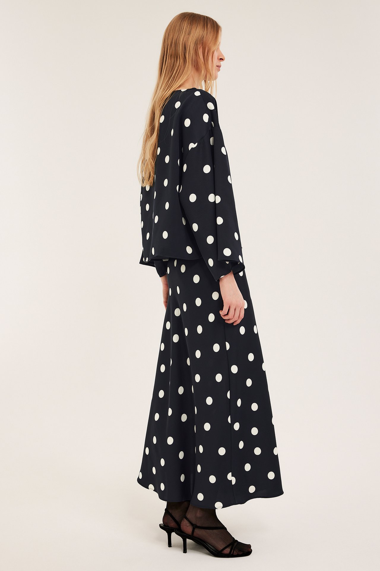 Widok z boku: Kobieta w czarnej bluzce satynowej w kropki, szerokie rękawy, spódnica i czarne obcasy.