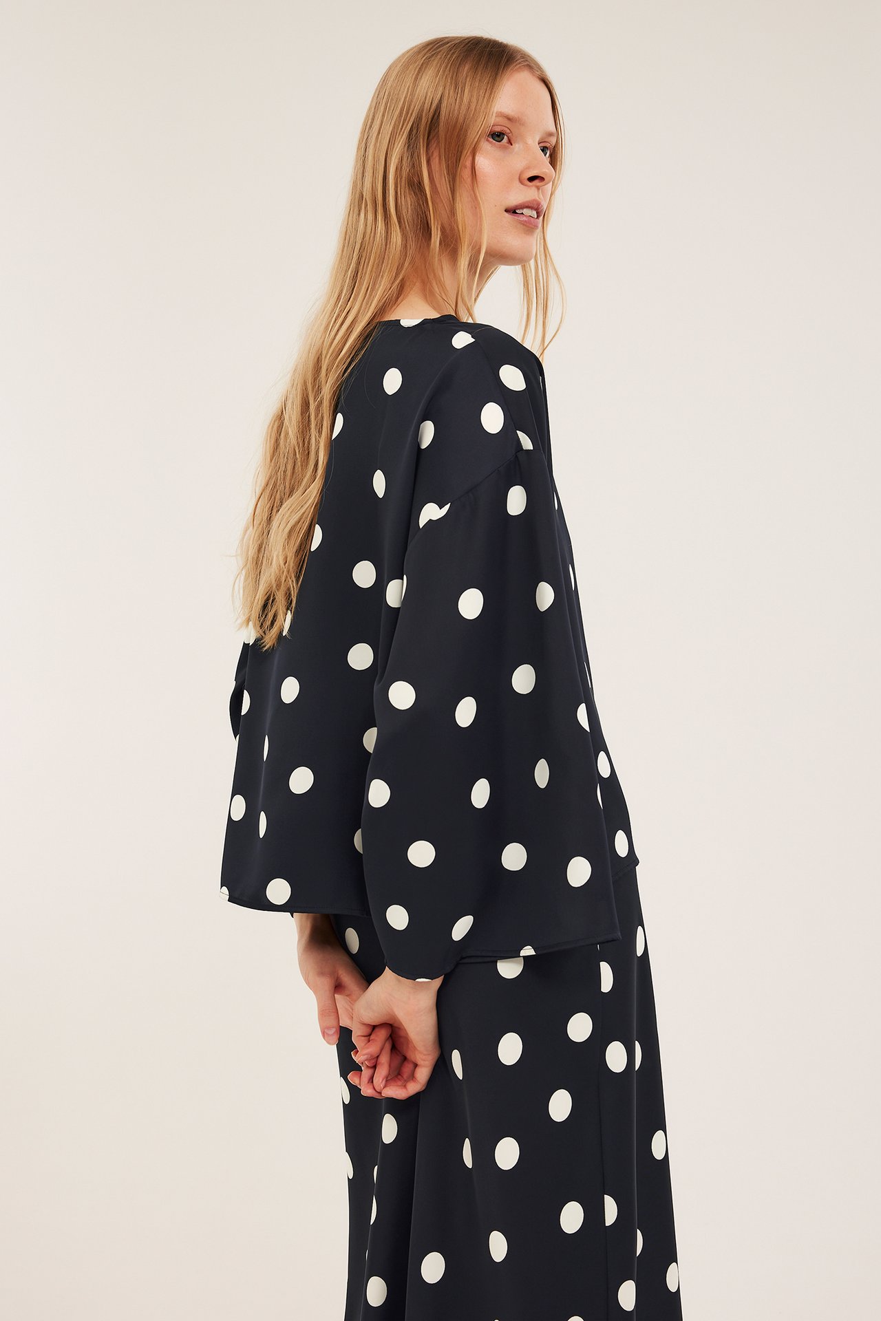 Widok z boku kobiety w czarnej bluzce satynowej w kropki z szerokimi rękawami i pasującej spódnicy.