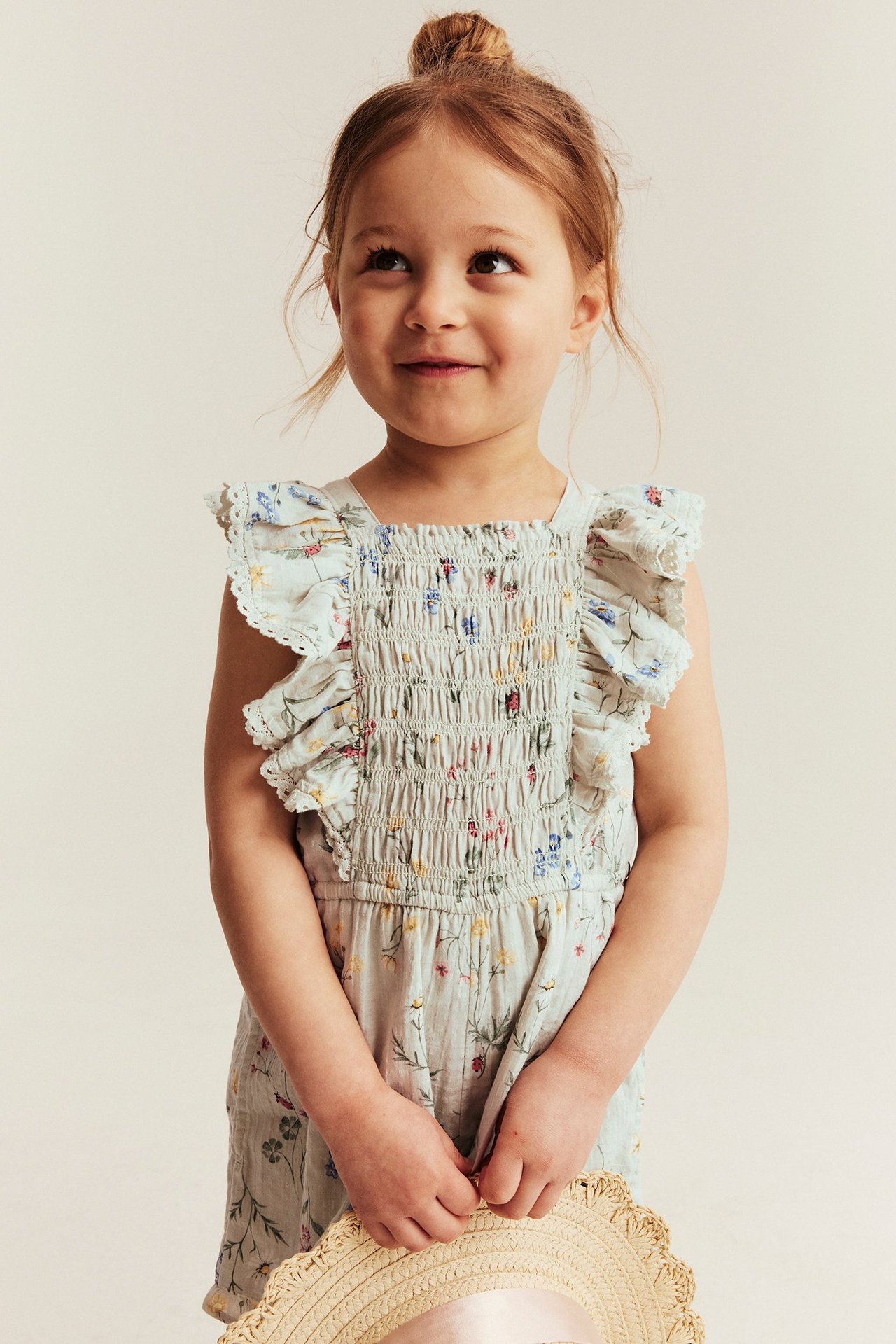 Edestä näkymä lapsesta, yllään vaaleanvihreä kukkakuvioinen smockattu haalari röyhelöillä, pitelee olkihattua.