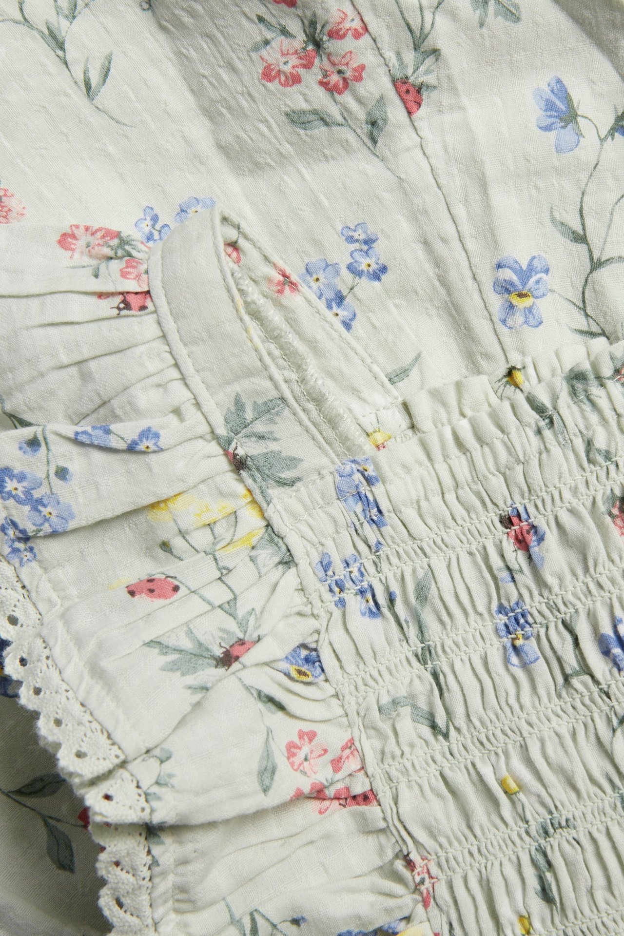 Närbild av en ljusgrön blommig vävd jumpsuit med smockresår, volanger och spetskant.