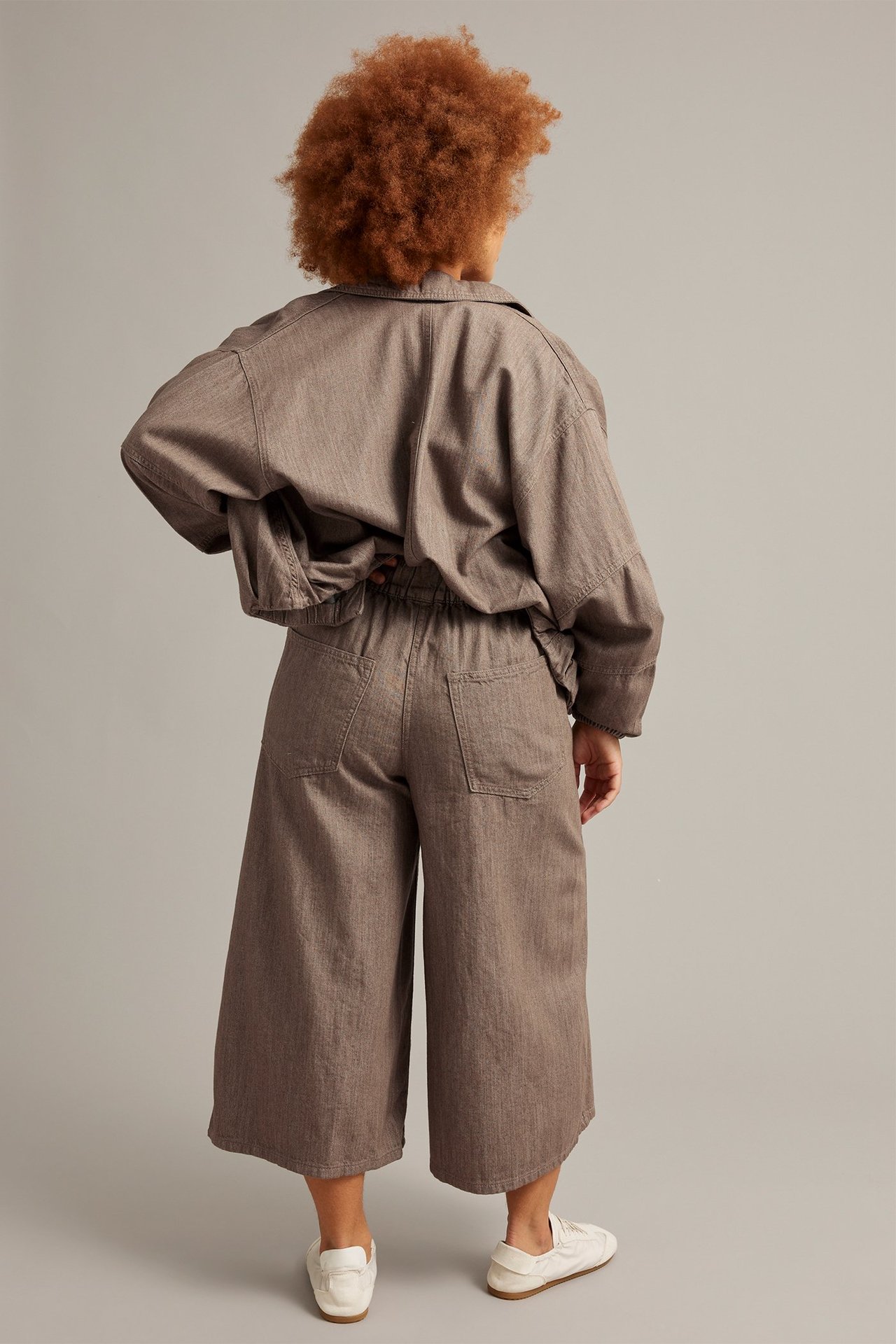 Bakfra av kvinne i brune melange bomullsculottejeans, matchende jakke og hvite joggesko.