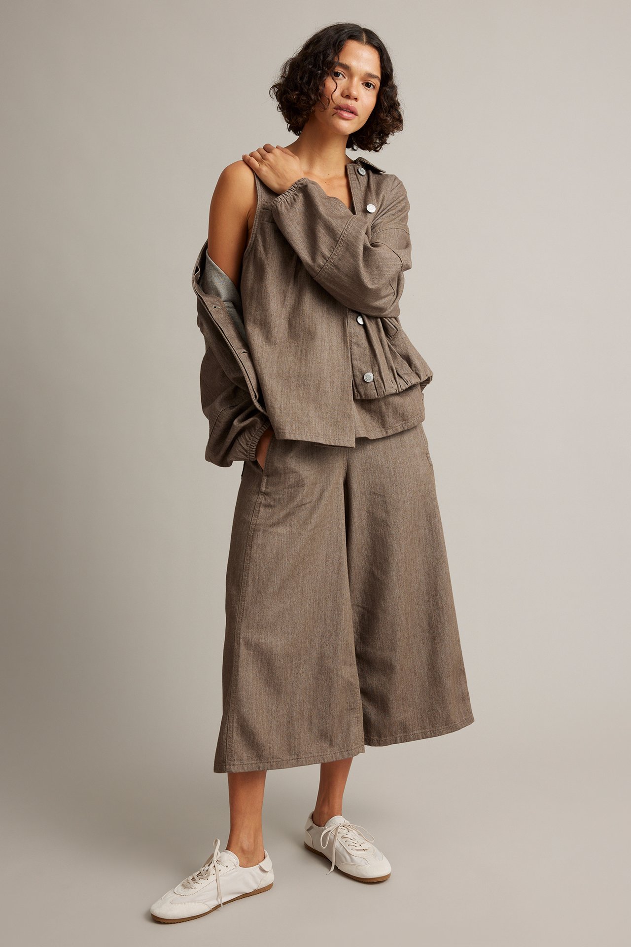 Front view: Kvinna i brun texturerad jacka, matchande linne, culottes och vita sneakers.