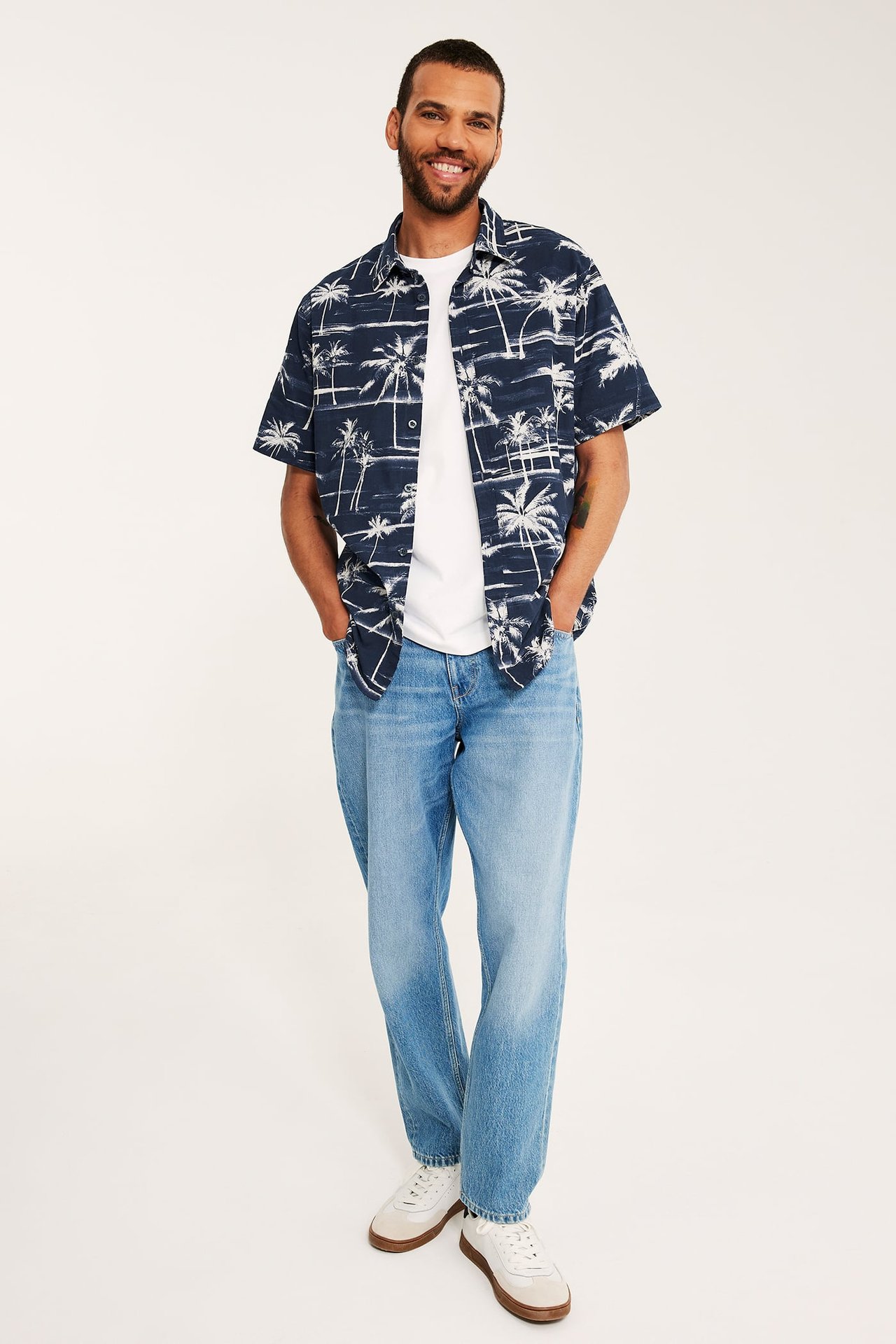 Frontvy. Man i mörkblå mönstrad kortärmad skjorta, vit t-shirt, ljusblå jeans och vita sneakers.