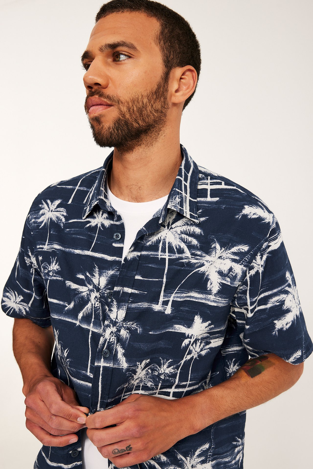 Man knäpper upp mörkblå kortärmad skjorta med vitt palmträdsmönster, buren över vit t-shirt.