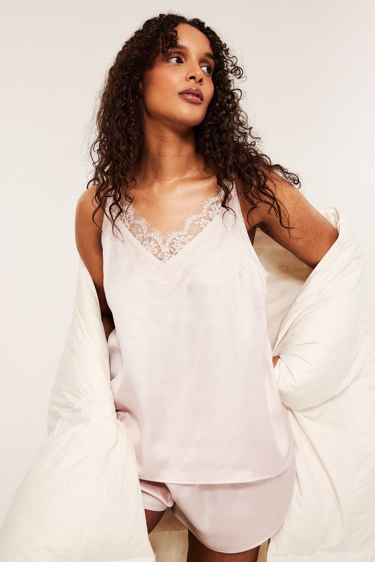 Framifrån: Kvinna i rosa satinpyjamas med spetskantad camisole och shorts, draperad i ett gräddvitt täcke.