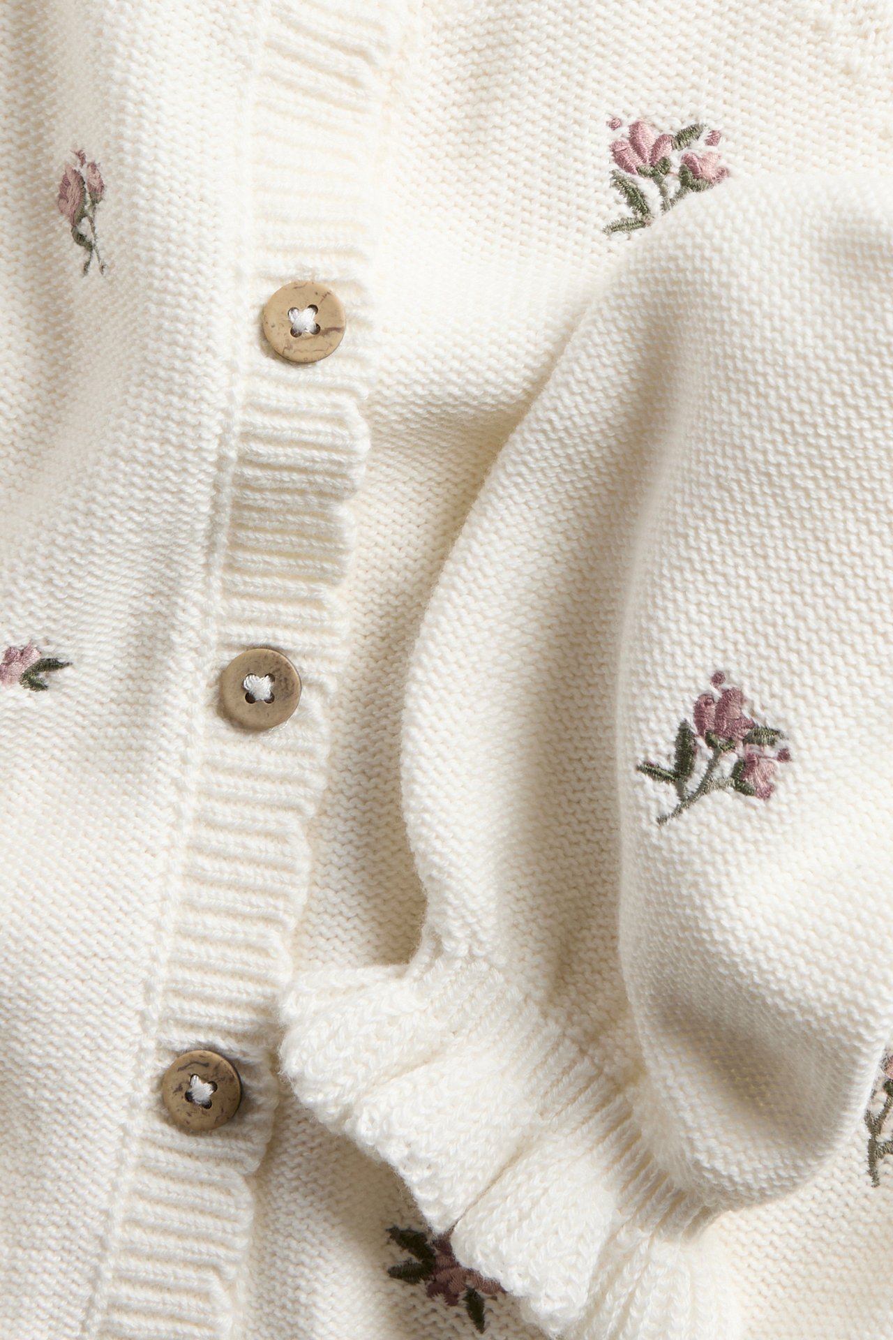 Nærbilde offwhite strikket bomullskardigan med broderte rosa blomster, treknapper og volangmudd.