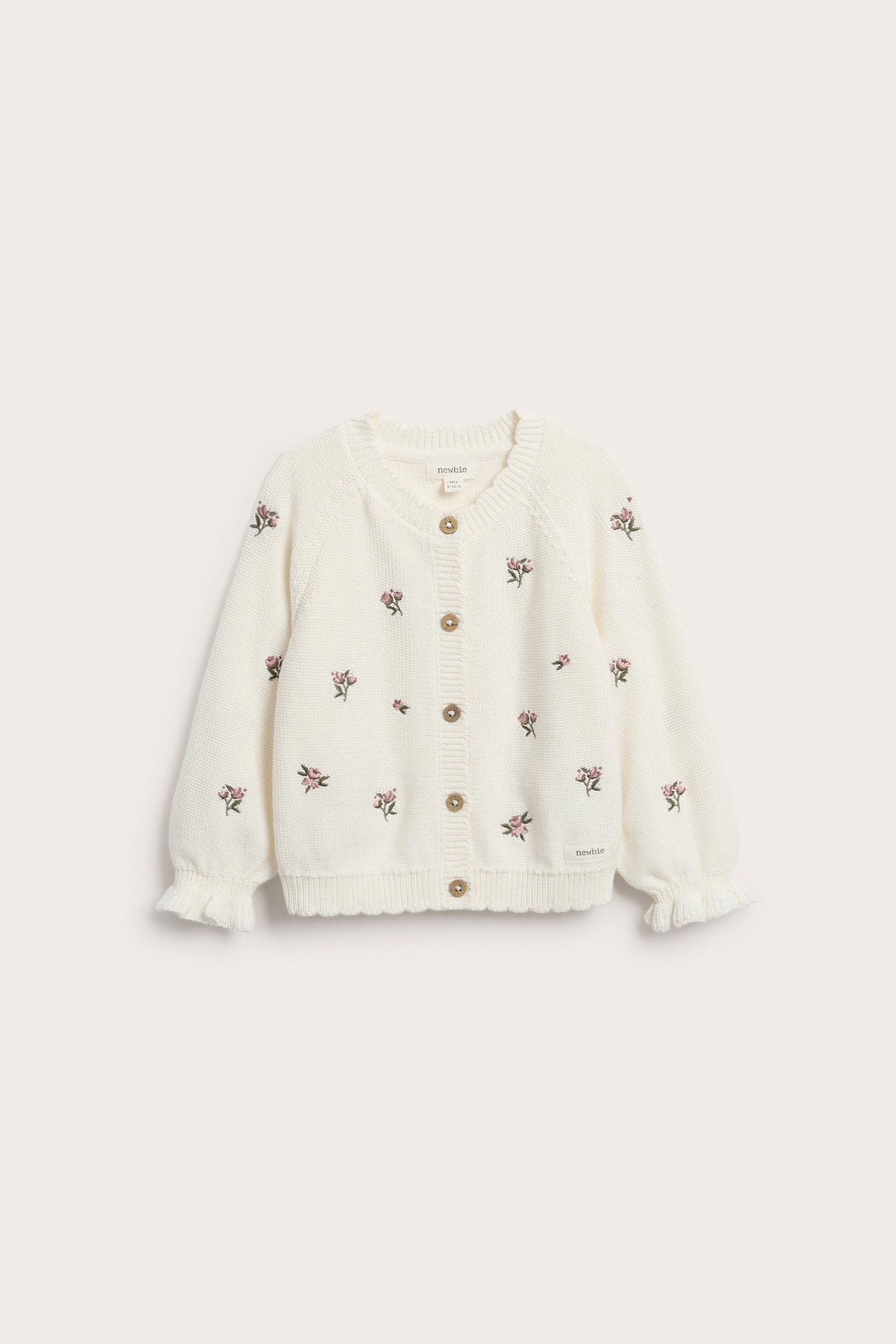 Frontbilde: off-white strikket barnecardigan i bomull med broderte rosa blomster, knapper, kamskjellkant og rysjede mansjetter.