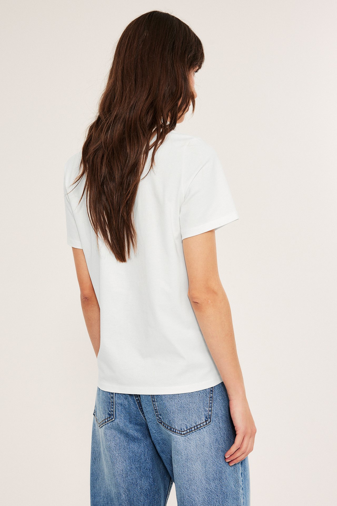 Kvinna i vit t-shirt med rak passform och blåjeans, sida på, bakgrund vit.