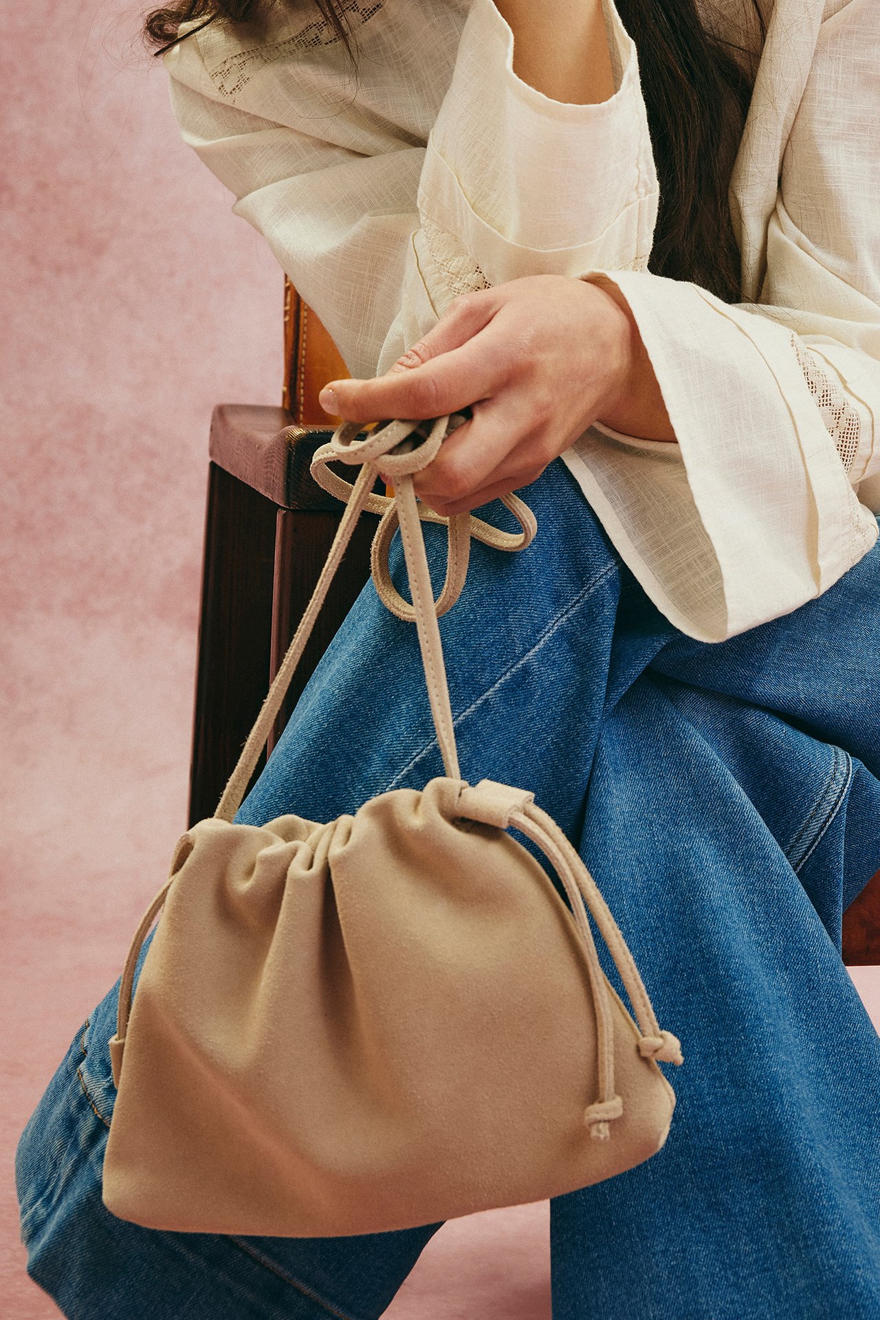 Närbild: kvinna håller beige mocka axelremsväska med dragsko, bär vit blus, blå jeans.
