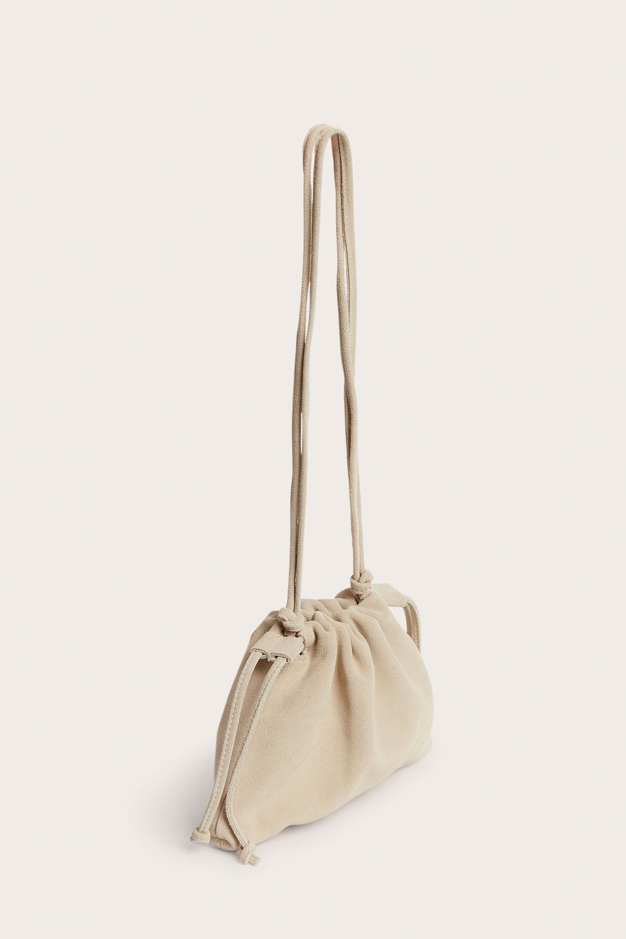 Skośny widok z przodu małej beżowej zamszowej torebki worka ze ściągaczem i regulowanym paskiem.