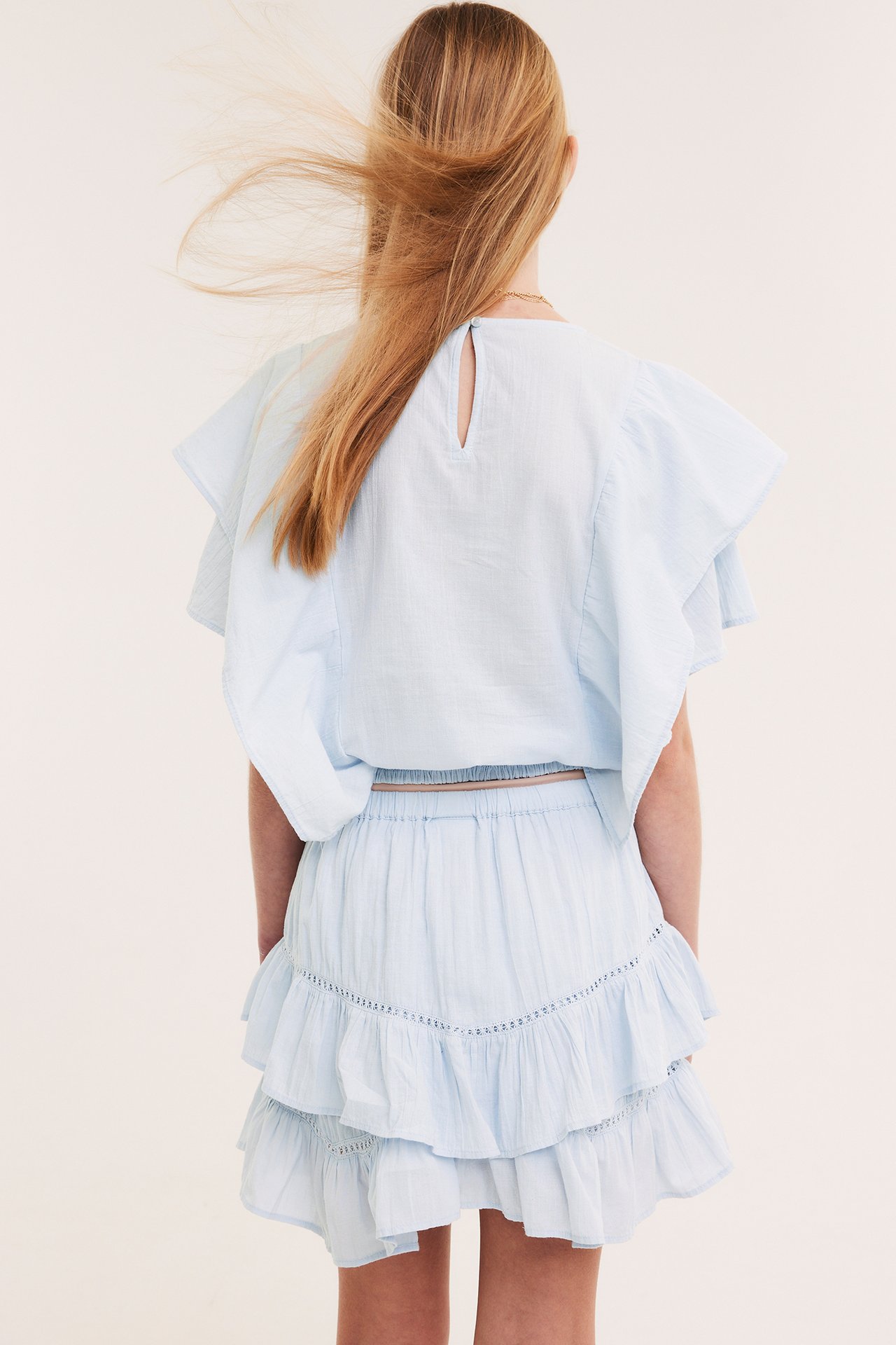 Lapsi takaa päin vaaleansinisessä puuvillapuserossa, jossa röyhelöitä, ja yhteensopivassa laskostetussa hameessa.