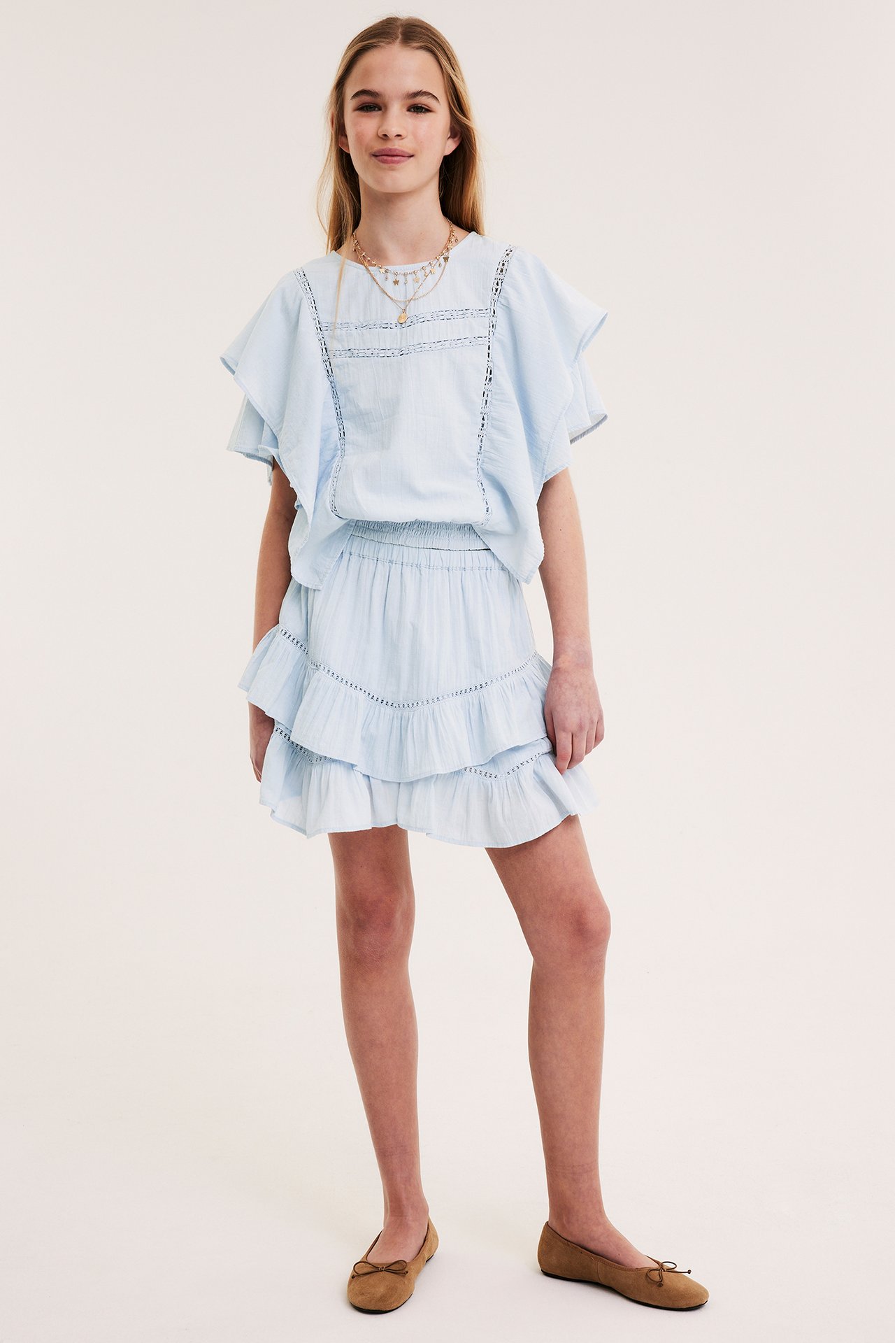 Lapsi yllään vaaleansininen puuvillainen batwing-pusero pitsillä, smokkiryppyvyötäröllä, yhteensopiva hame ja ruskeat ballerinat.