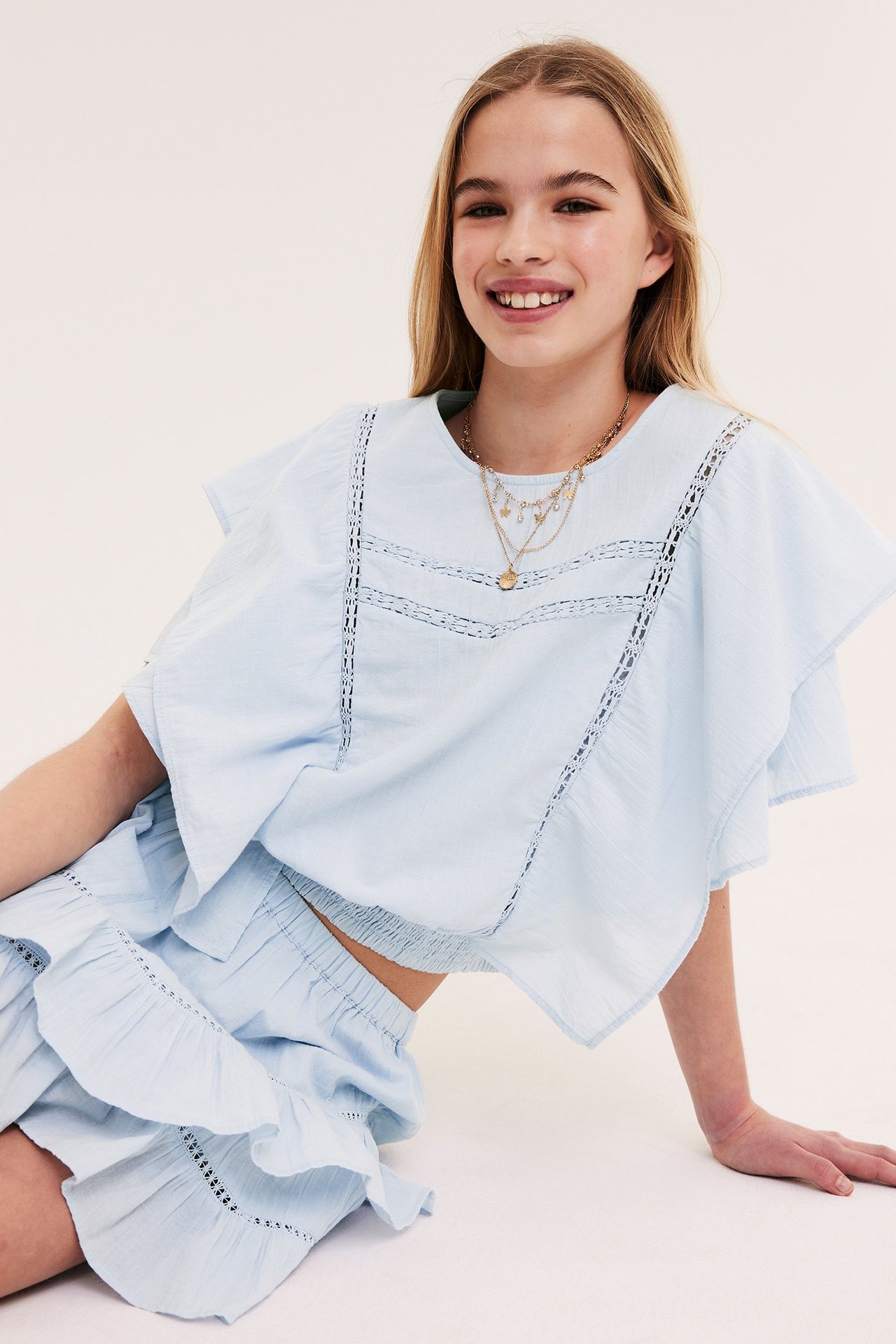 Framifrån: Barn i ljusblå bomullsblus med fladdermusärm, spets, smock i midjan, matchande kjol, halsband.