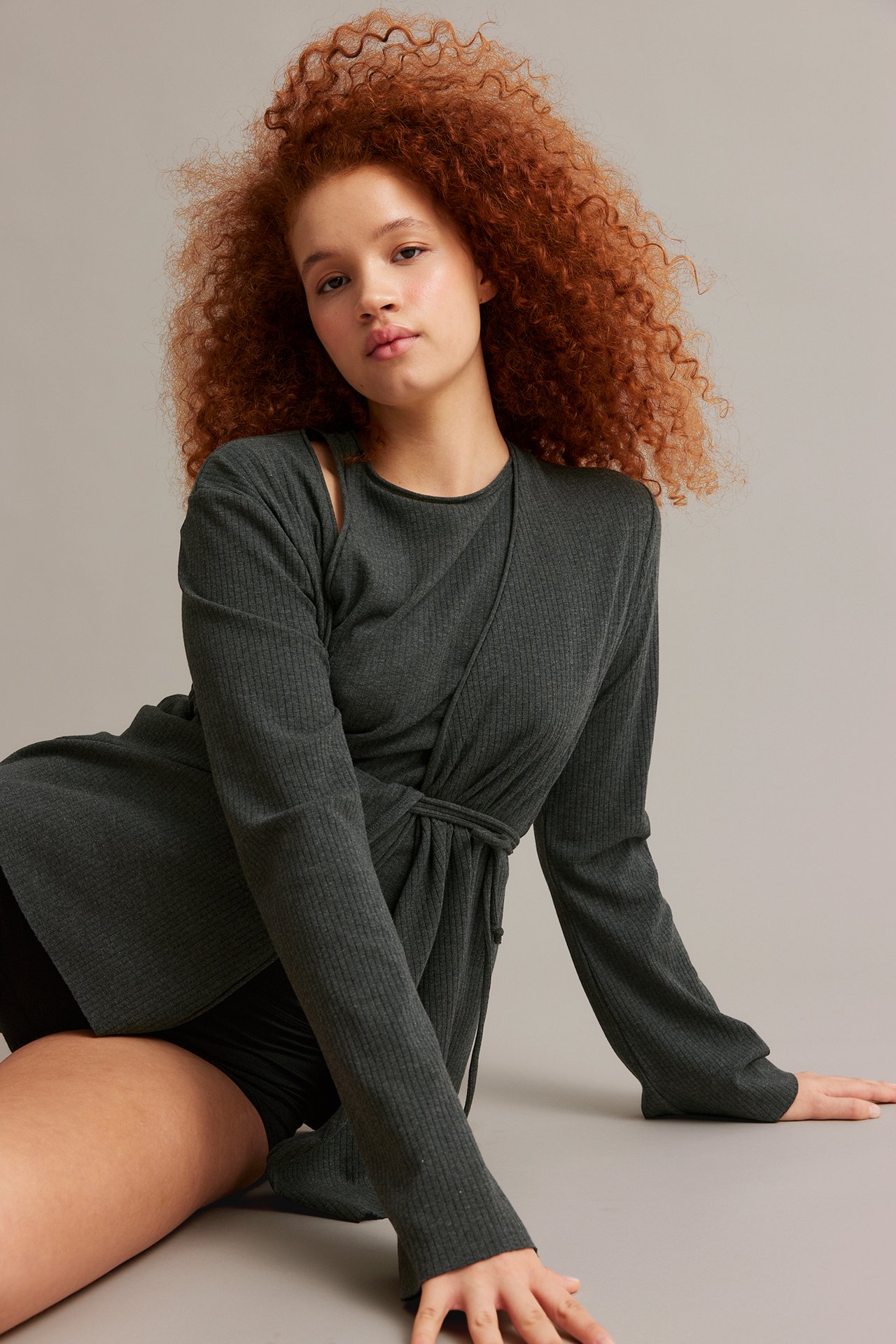 Widok z przodu: Kobieta w ciemnoszarym melanżowym prążkowanym kardiganie wiązanym w talii i czarnych szortach.