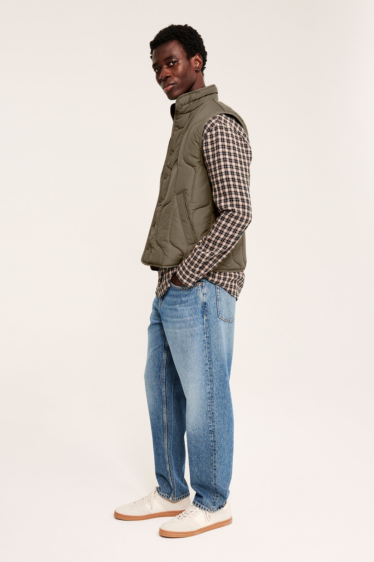 Sidovy av man i khaki-grön quiltad väst, rutig skjorta, blå jeans och vita sneakers.