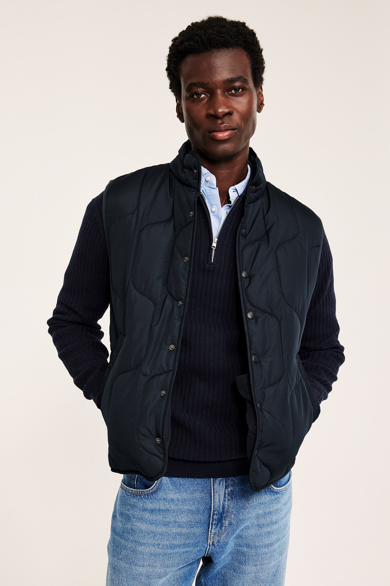 Framifrån: Man bär mörkblå quiltad väst med tryckknappar, över mörk tröja och blå jeans.