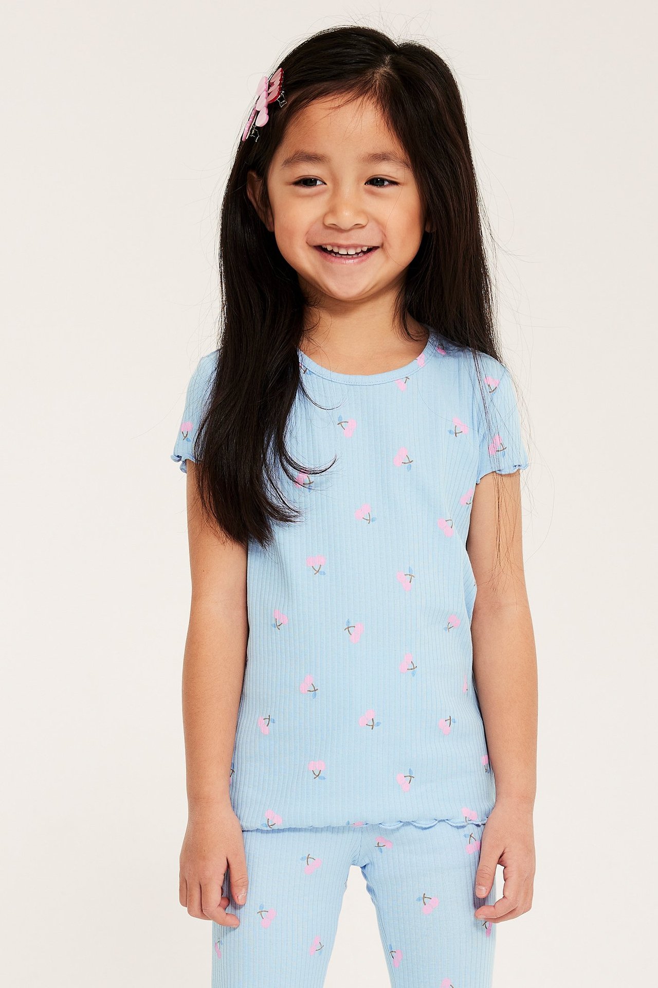 Uśmiechnięte dziecko w jasnoniebieskim topie w wiśnie, pasujących legginsach i różowych spinkach, widok z przodu.