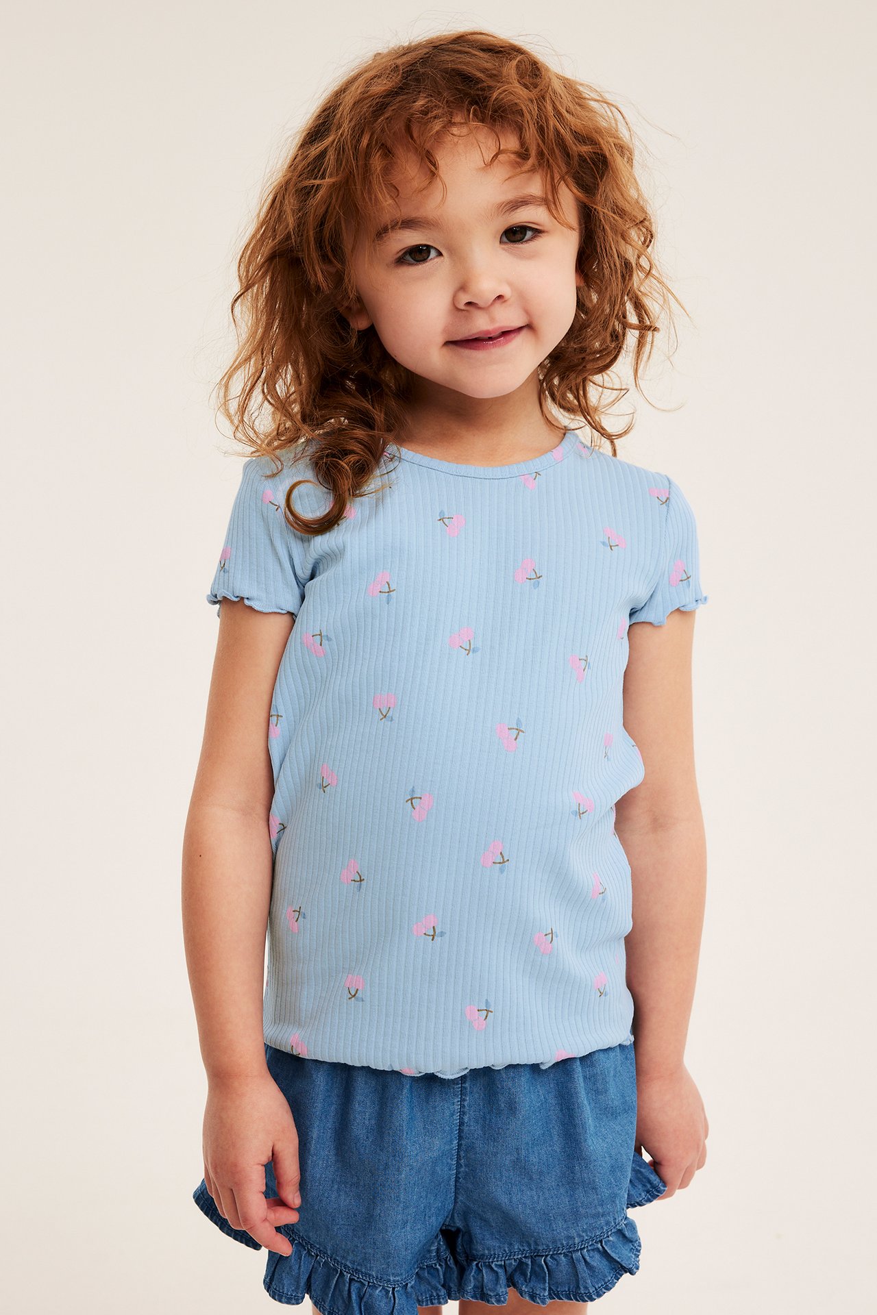 Przód: dziecko w jasnoniebieskim prążkowanym topie w wiśnie i niebieskich jeansowych szortach z falbanami.