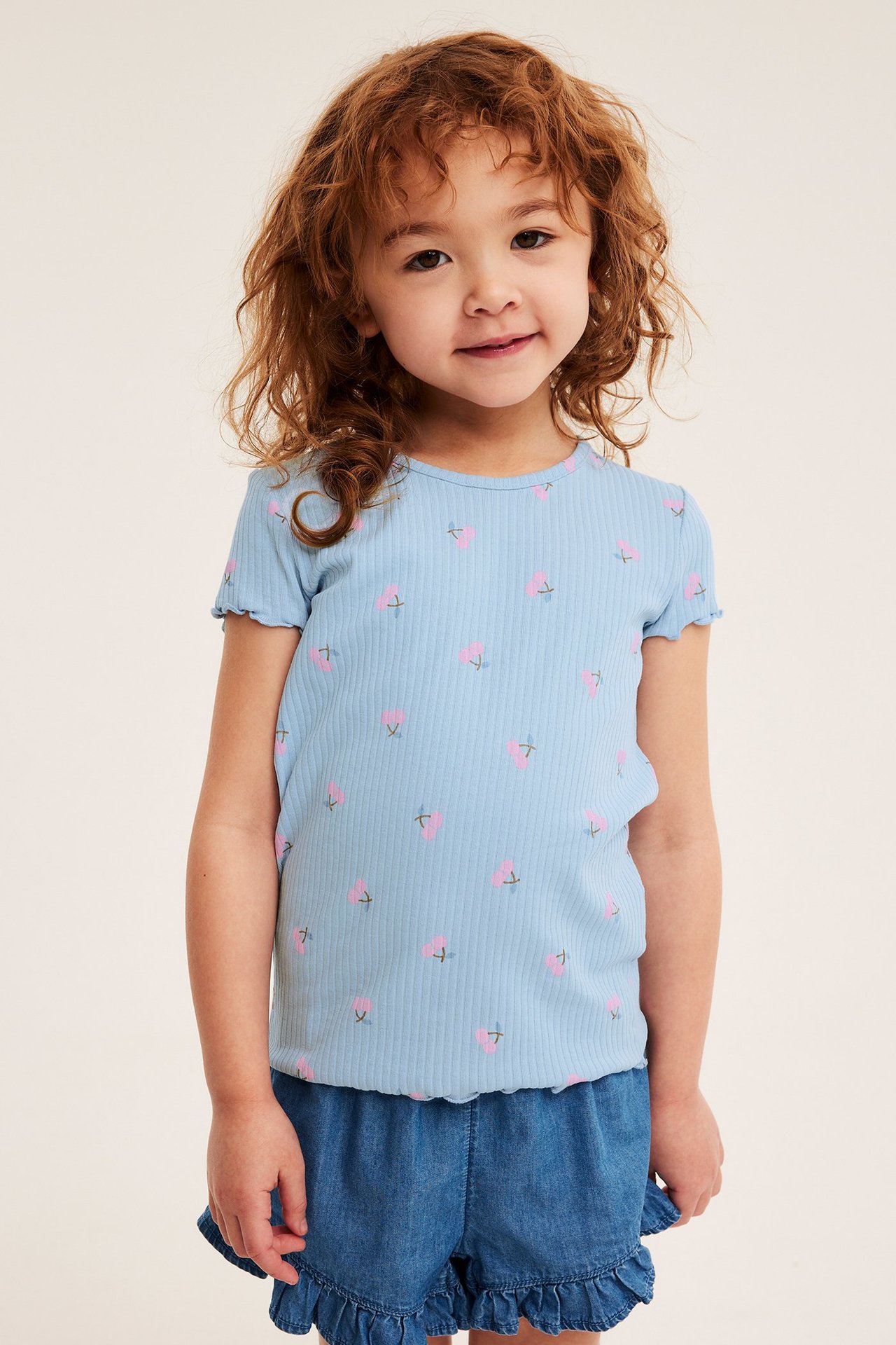 Edestä: lapsi vaaleansinisessä ribbitopissa kirsikkaprintillä ja sinisissä rypytetyissä farkkushortseissa.