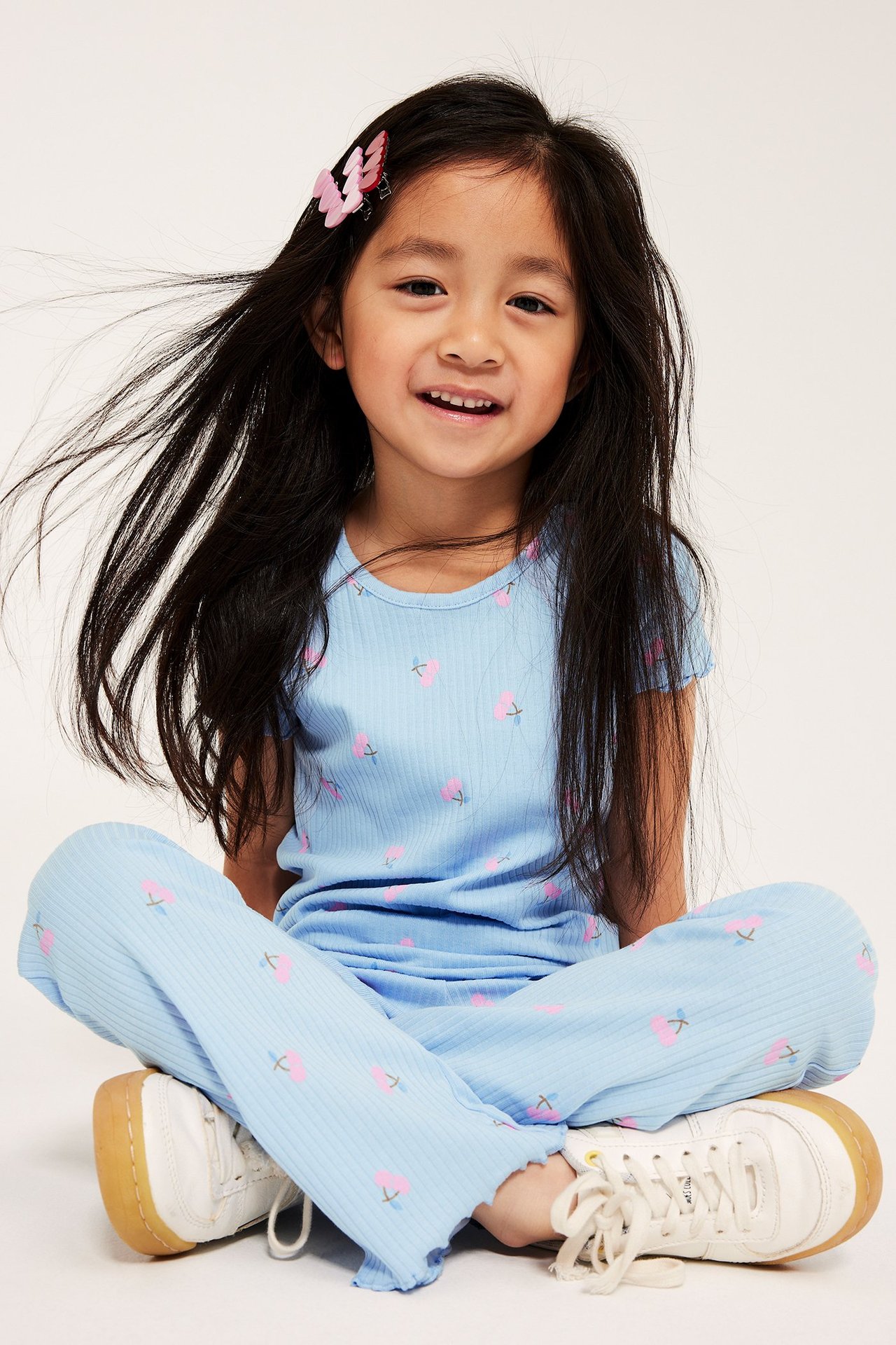Lapsi vaaleansinisessä ribbitopissa kirsikkaprintillä, yhteensopivat housut, valkoiset tennarit ja pinkit perhosklipsit.