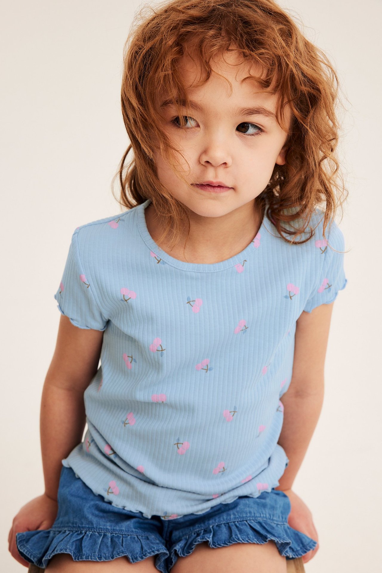 Frontvy av ett barn i ljusblå ribbad topp med rosa körsbärstryck och blå volangshorts i denim.