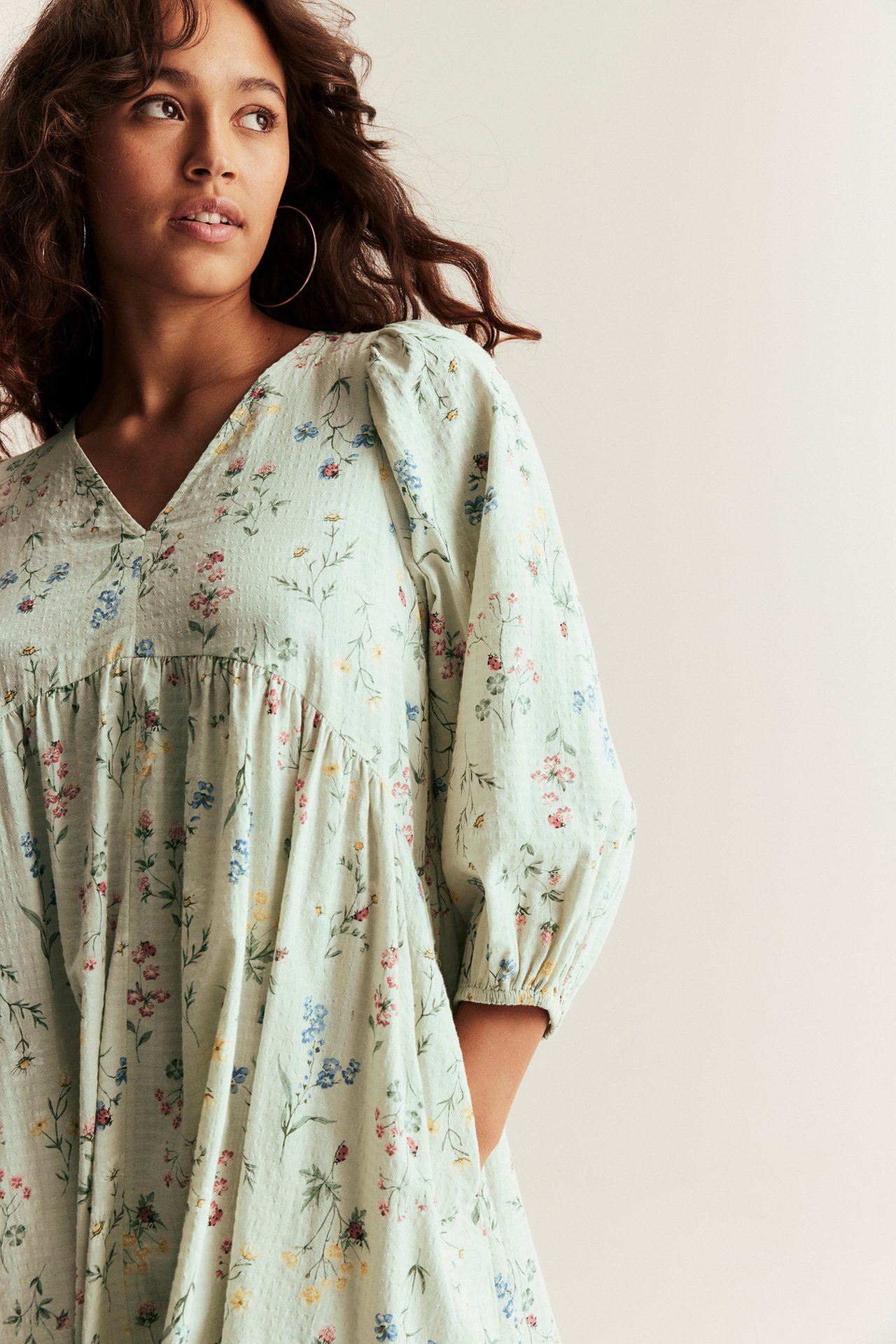 Nainen yllään vaaleanvihreä kukkakuvioinen puuvillamekko, jossa v-pääntie, pöyhkeät hihat ja vannekorvakorut.