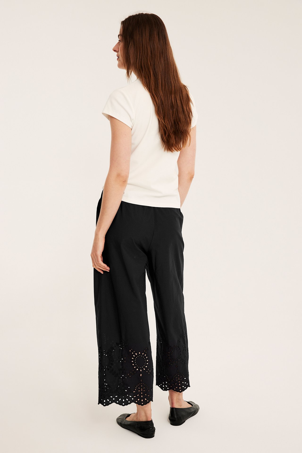 Naisen takasivukuva: mustat leveät broderie anglaise -puuvillahousut, kermanvärinen toppi, mustat kengät.