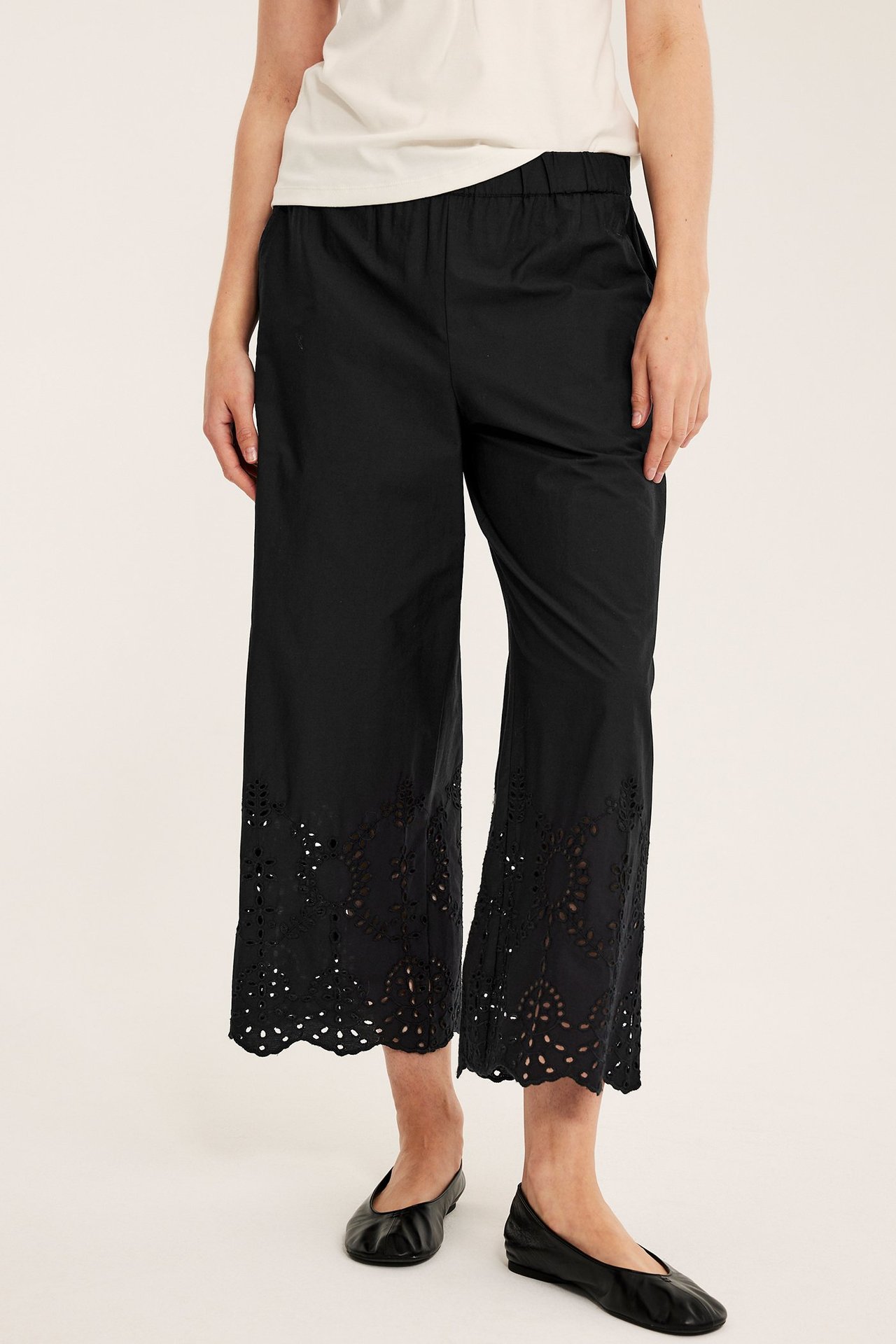 Edestäpäin: nainen mustissa brodeeratuissa puuvillapopliinicullottehousuissa, valkoinen t-paita, mustat kengät.