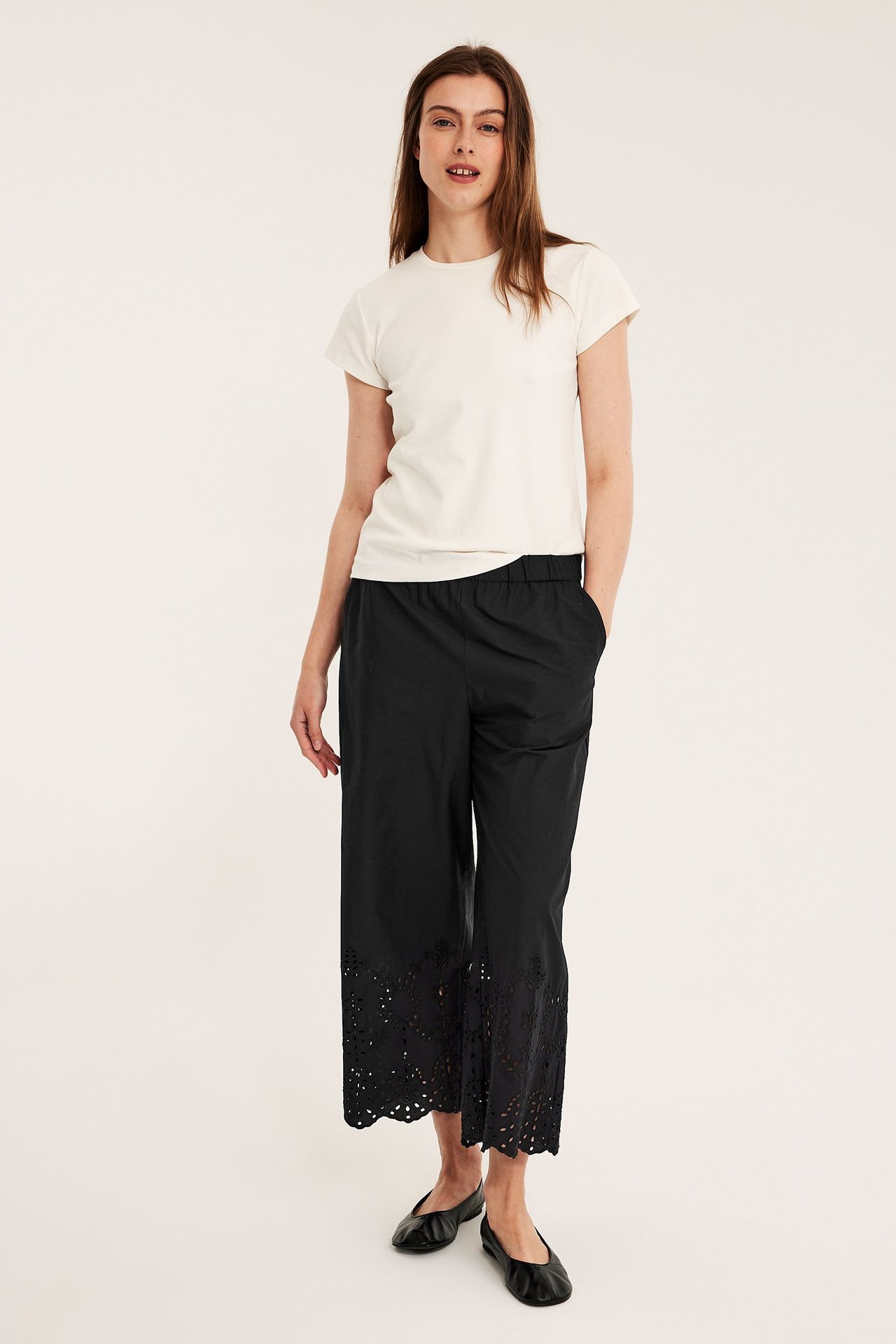 Naisen edestä: mustat pohjepituiset puuvillapopliinihousut englantilaiskirjonnalla, beige T-paita ja mustat ballerinat.