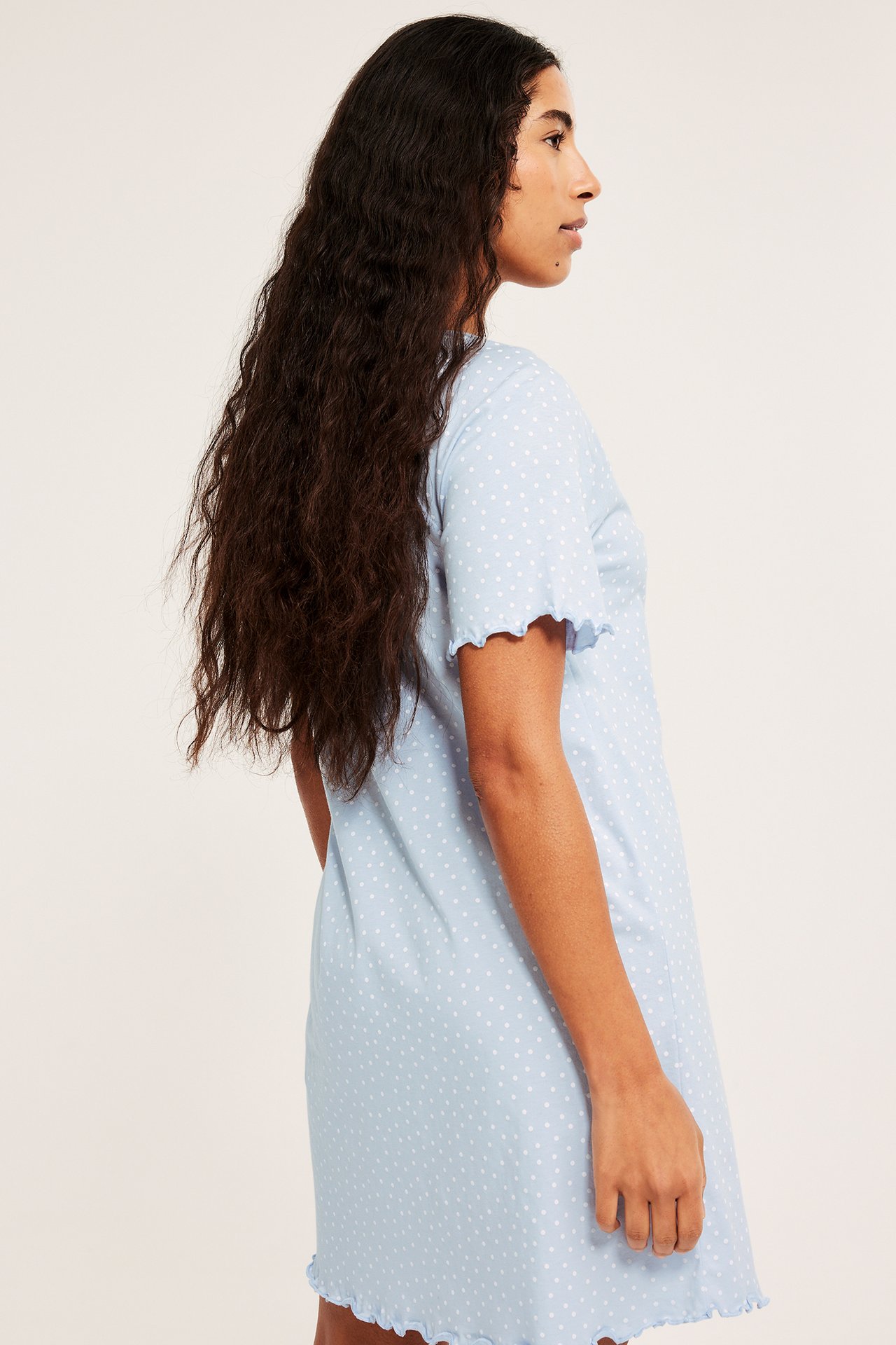 Widok z boku kobiety w jasnoniebieskiej, bawełnianej koszuli nocnej w białe kropki z falbanami.