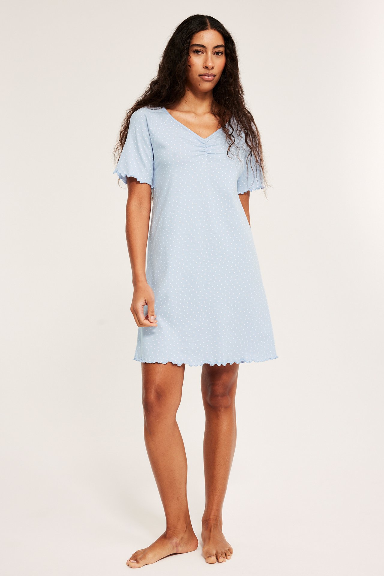 Widok z przodu: Kobieta w jasnoniebieskiej bawełnianej koszuli nocnej w kropki, z marszczonym dekoltem V i falistymi brzegami.