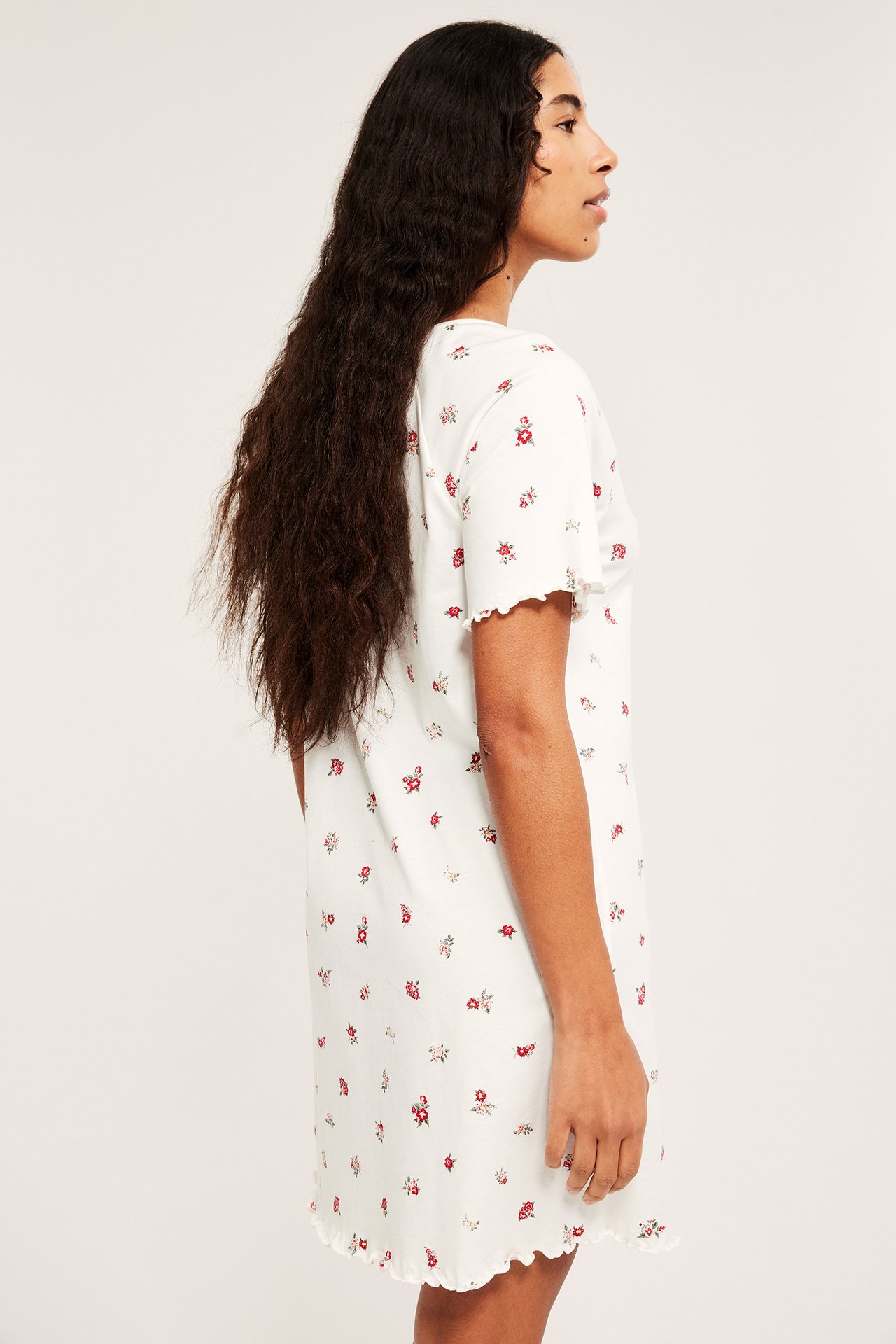 Sidebilde av en kvinne i et offwhite bomullsnattkjole med rødt blomstermønster og bølgete kanter.