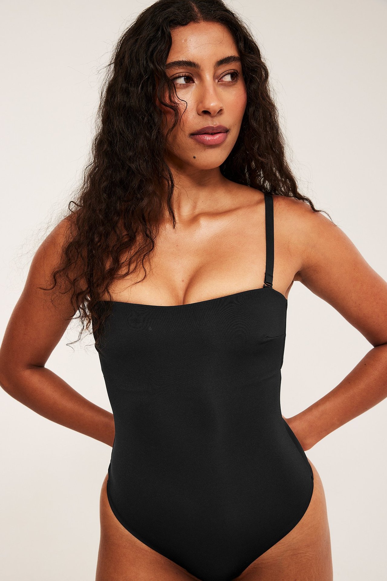 Frontbilde av kvinne i svart polstret stringbody med avtakbare stropper, laget av glatt, elastisk stoff.