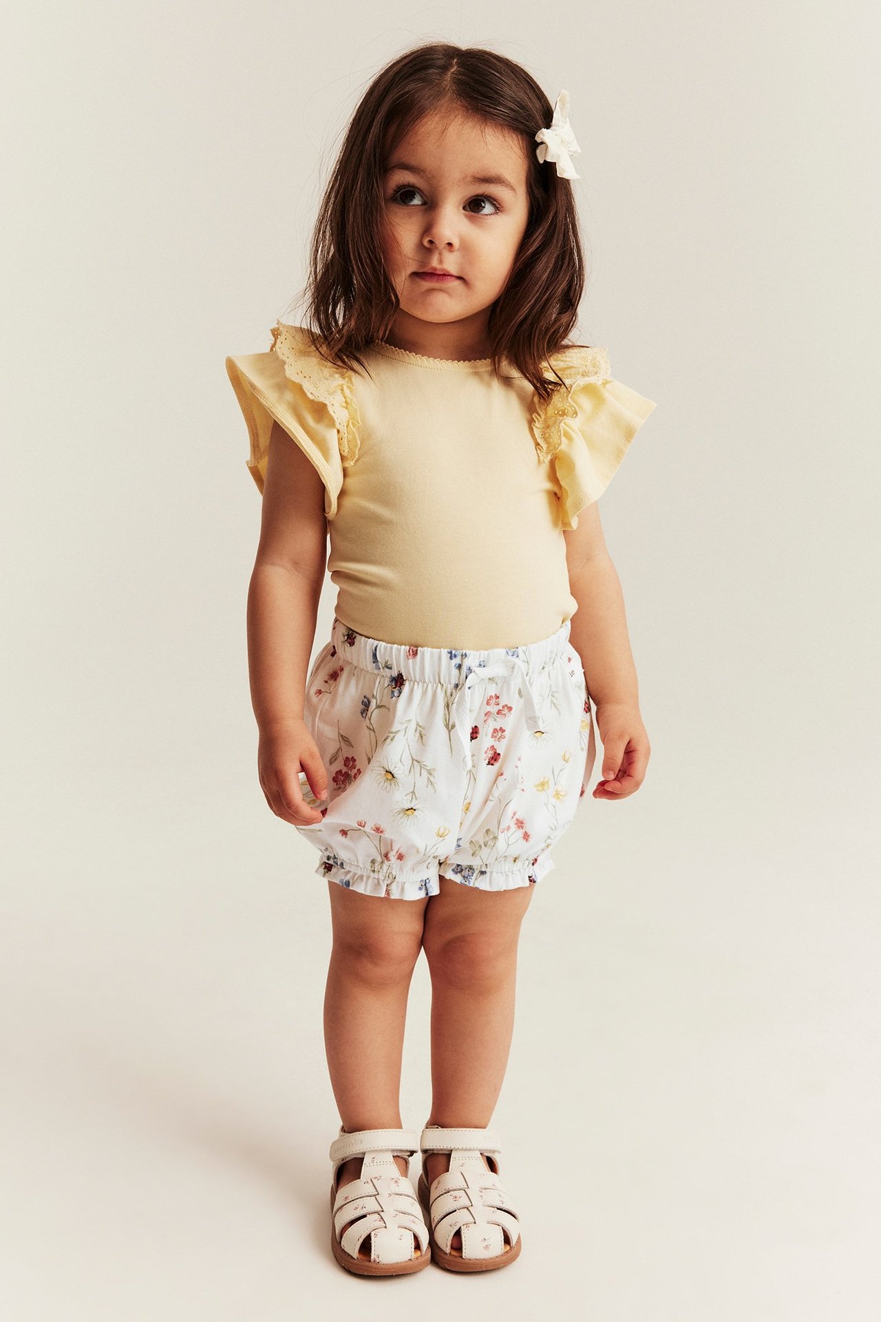 Frontbilde av et barn i hvite blomstrete jerseyshorts med volanger, en gul topp og lyse sandaler.