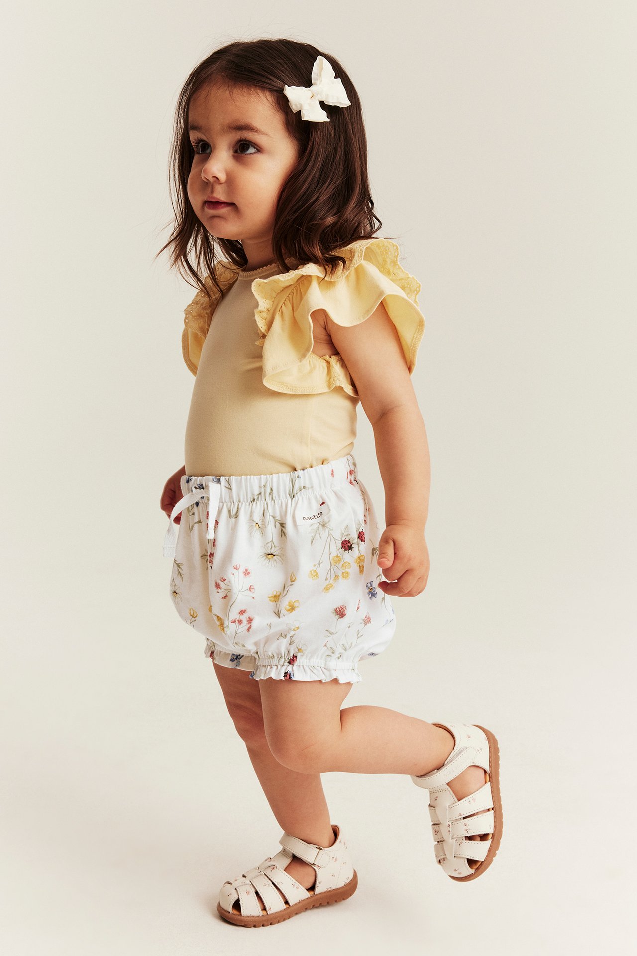 Vauvan sivukuva, yllään luonnonvalkoiset kukkakuvioiset pussi-shorts, keltainen röyhelöpaita ja valkoiset sandaalit.