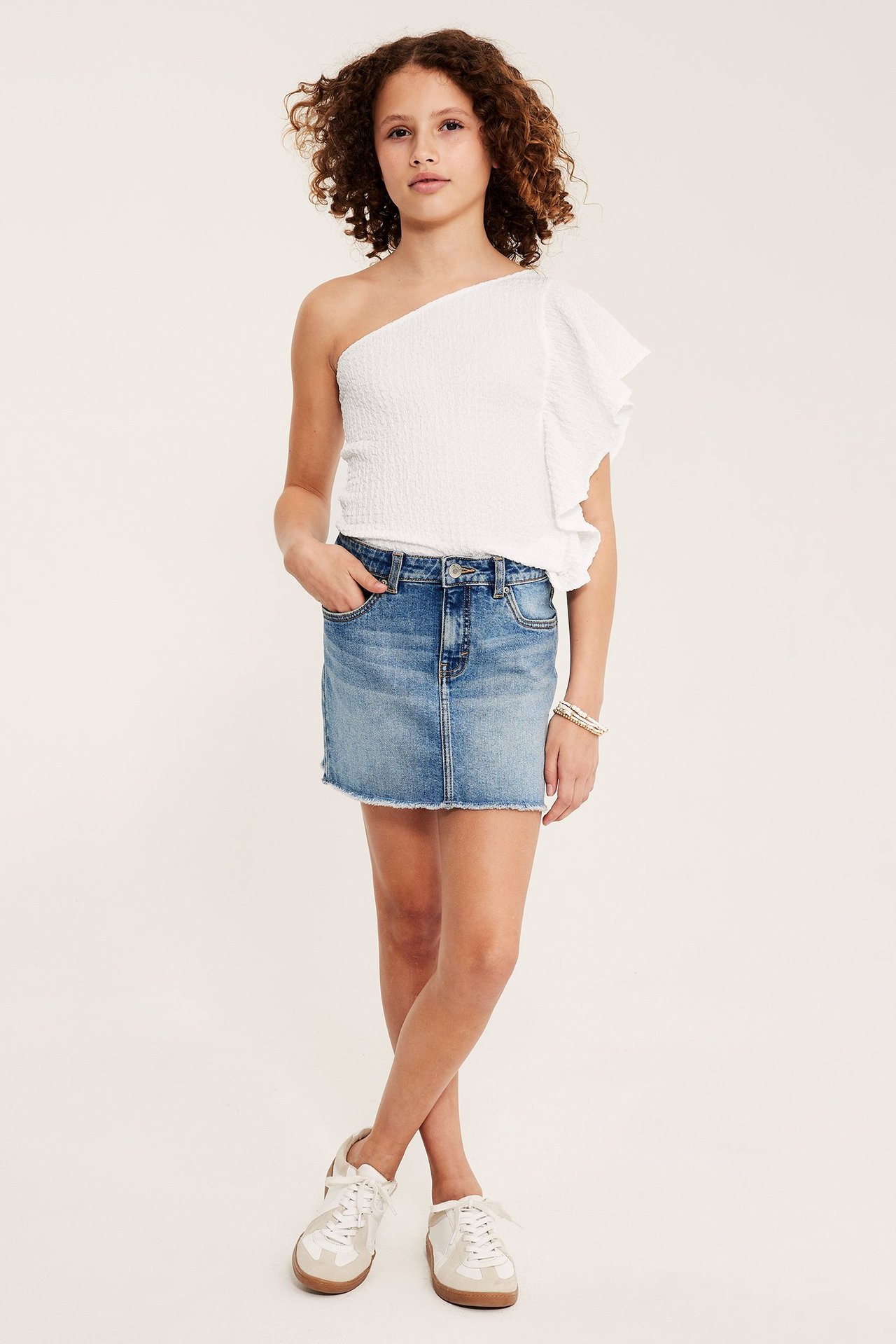 Framifrån en unge i vit smockad one shoulder-topp med volang, blå jeanskjol och vita sneakers.