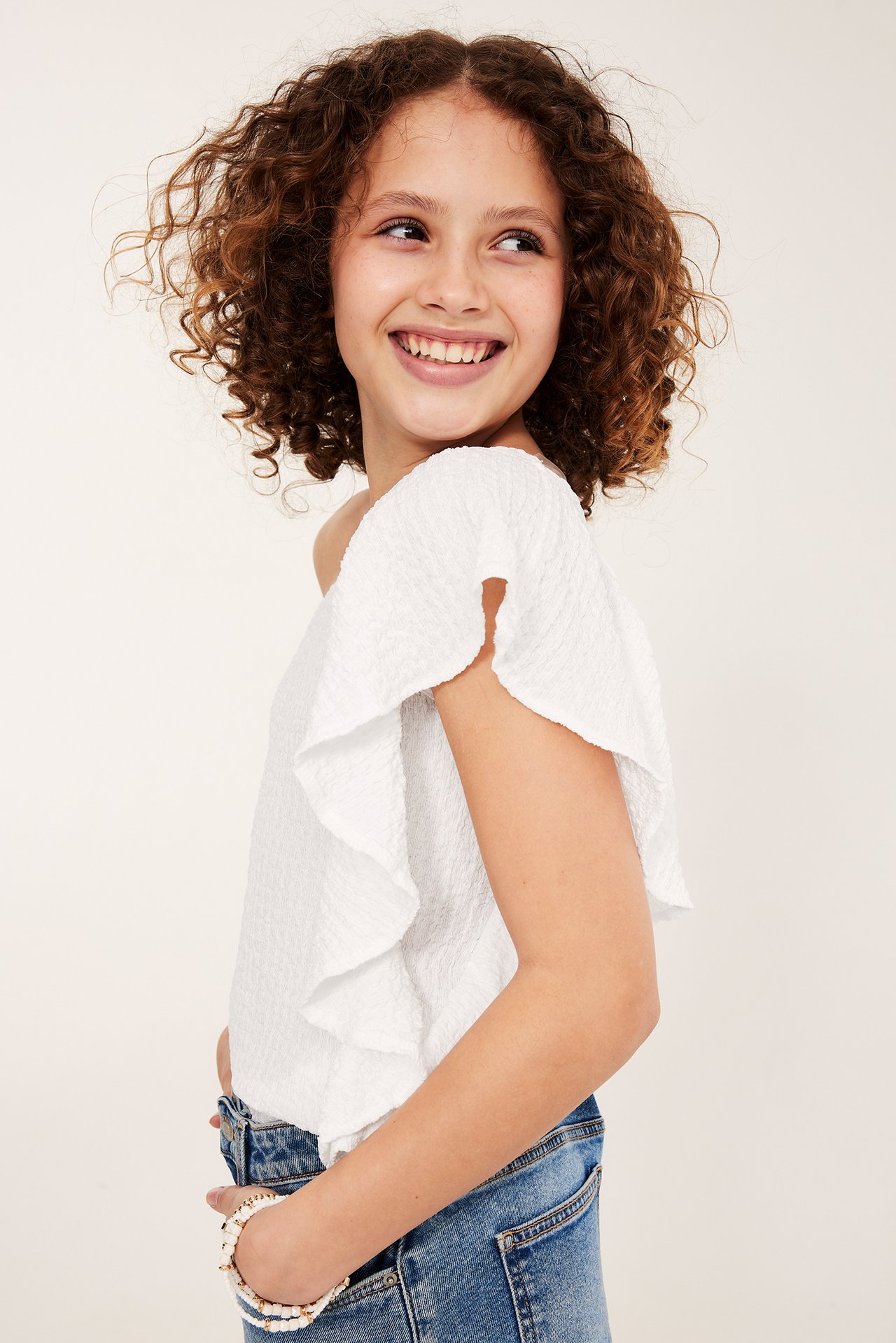 Sivukuva lapsesta, yllään valkoinen, toispuolinen röyhelötoppi, siniset farkut ja rannekoru.