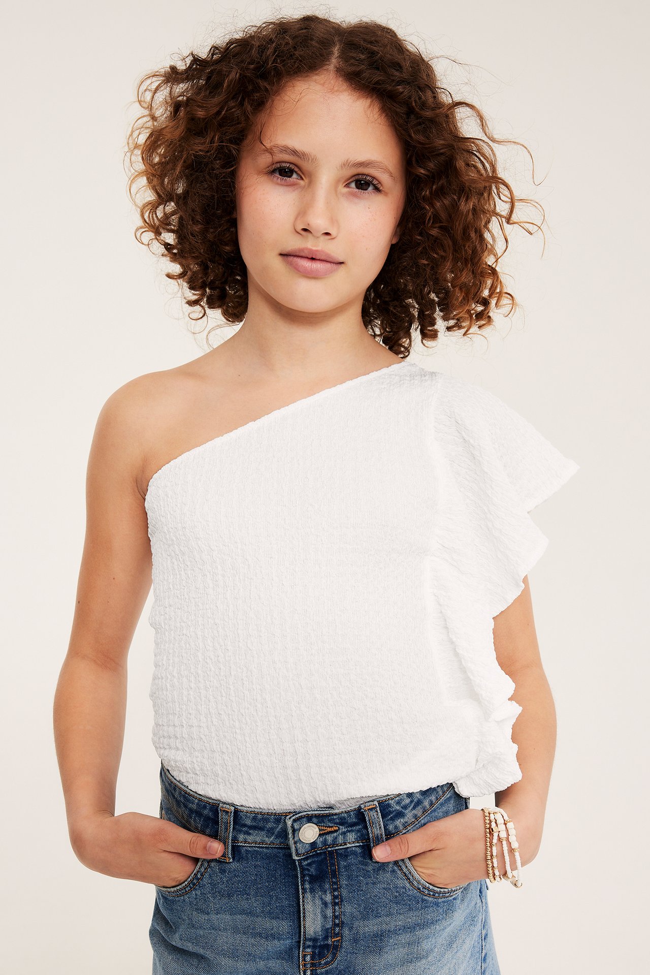 Widok z przodu: dziecko w białym topie na jedno ramię z falbaną, niebieskich jeansach i bransoletkach.