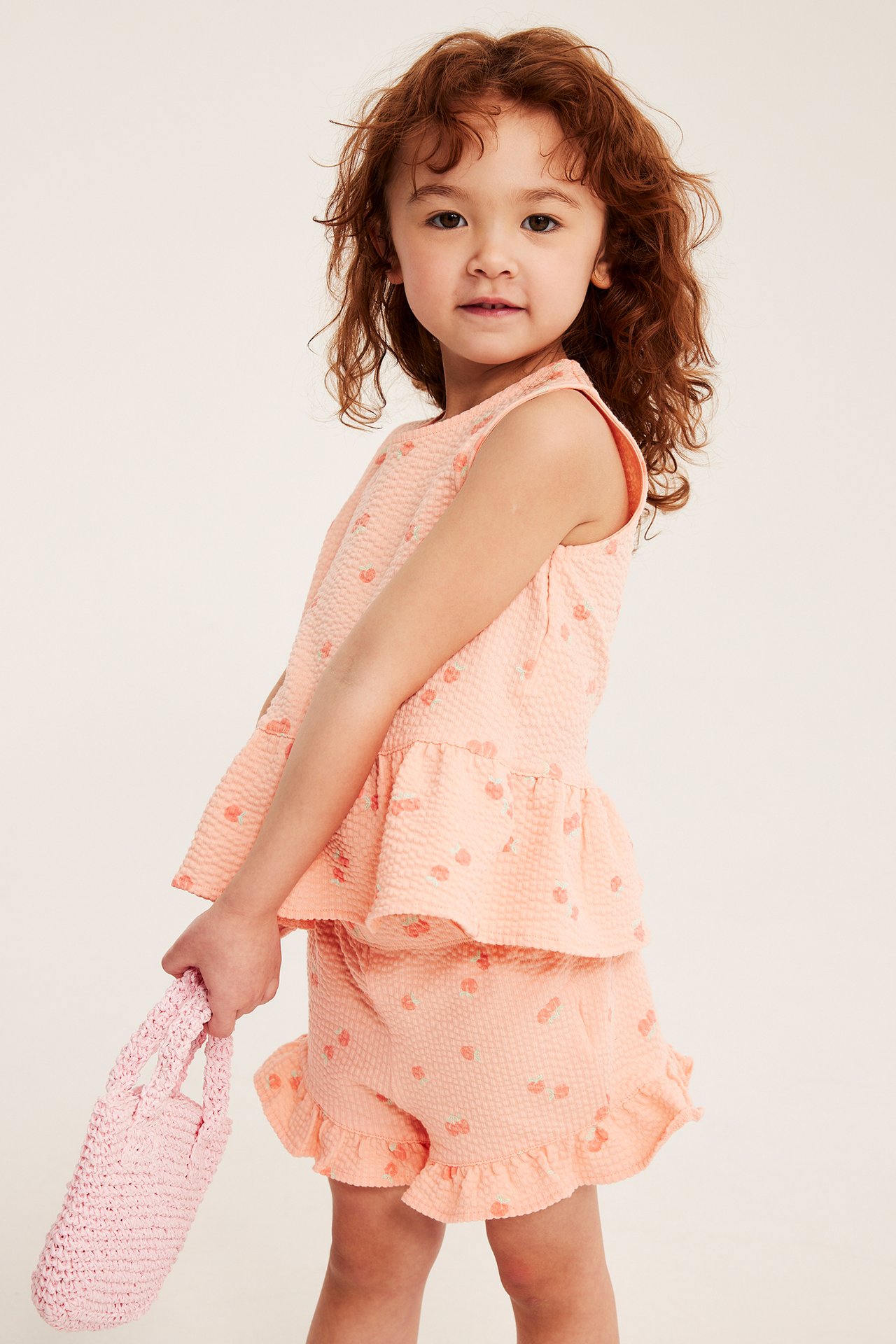 Widok z przodu dziecka w koralowych szortach z brzoskwiniowym wzorem i dopasowanym topie, trzymającego różową torebkę.
