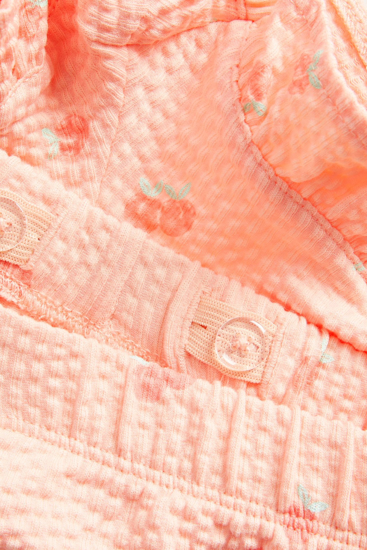 Zbliżenie na koralowe spodenki dziecięce z wzorem brzoskwiń, falbanką i regulowanym pasem.