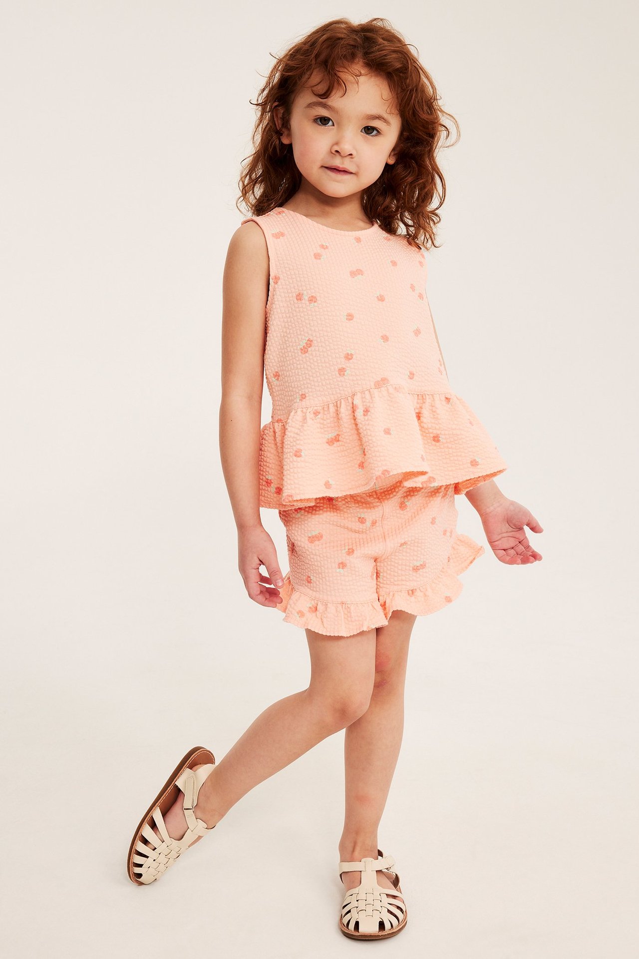 Z przodu, dziecko w koralowych szortach z wzorem brzoskwiń, falbanką, pasującym topie i beżowych sandałach.