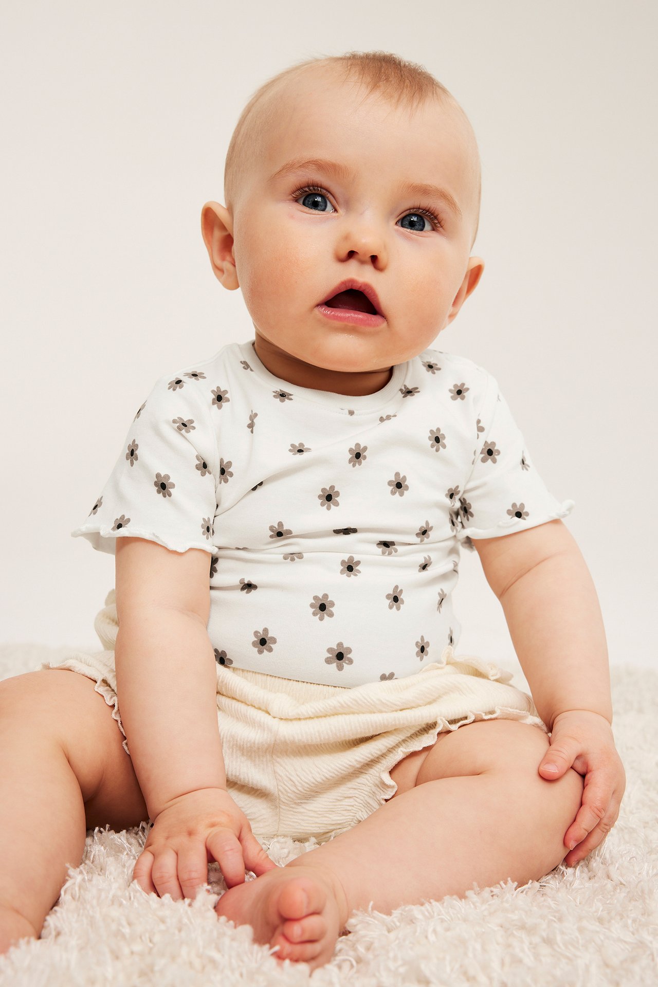 Vauva edestä: luonnonvalkoinen kukkakuvioinen lyhythihainen body ja vaaleat röyhelöshortsit.