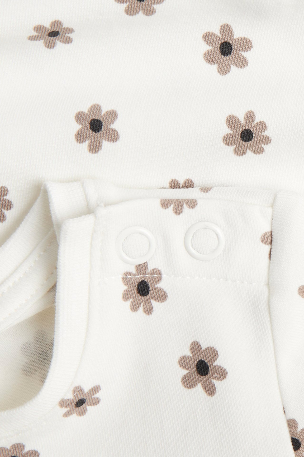 Närbild på en babys offwhite kortärmade bomullsbody med små brunt blommigt mönster och axelknappar.