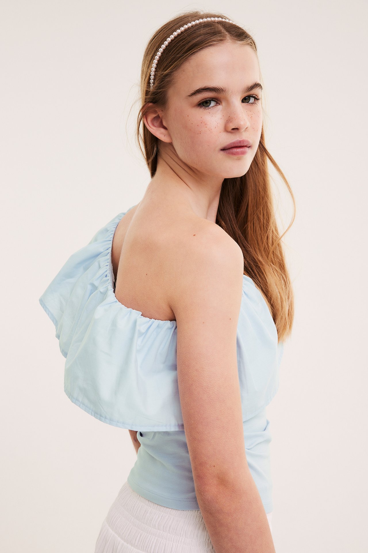 Sida vy av barn i ljusblå enaxlad volangtopp i bomull, vit kjol och pärlhårband.